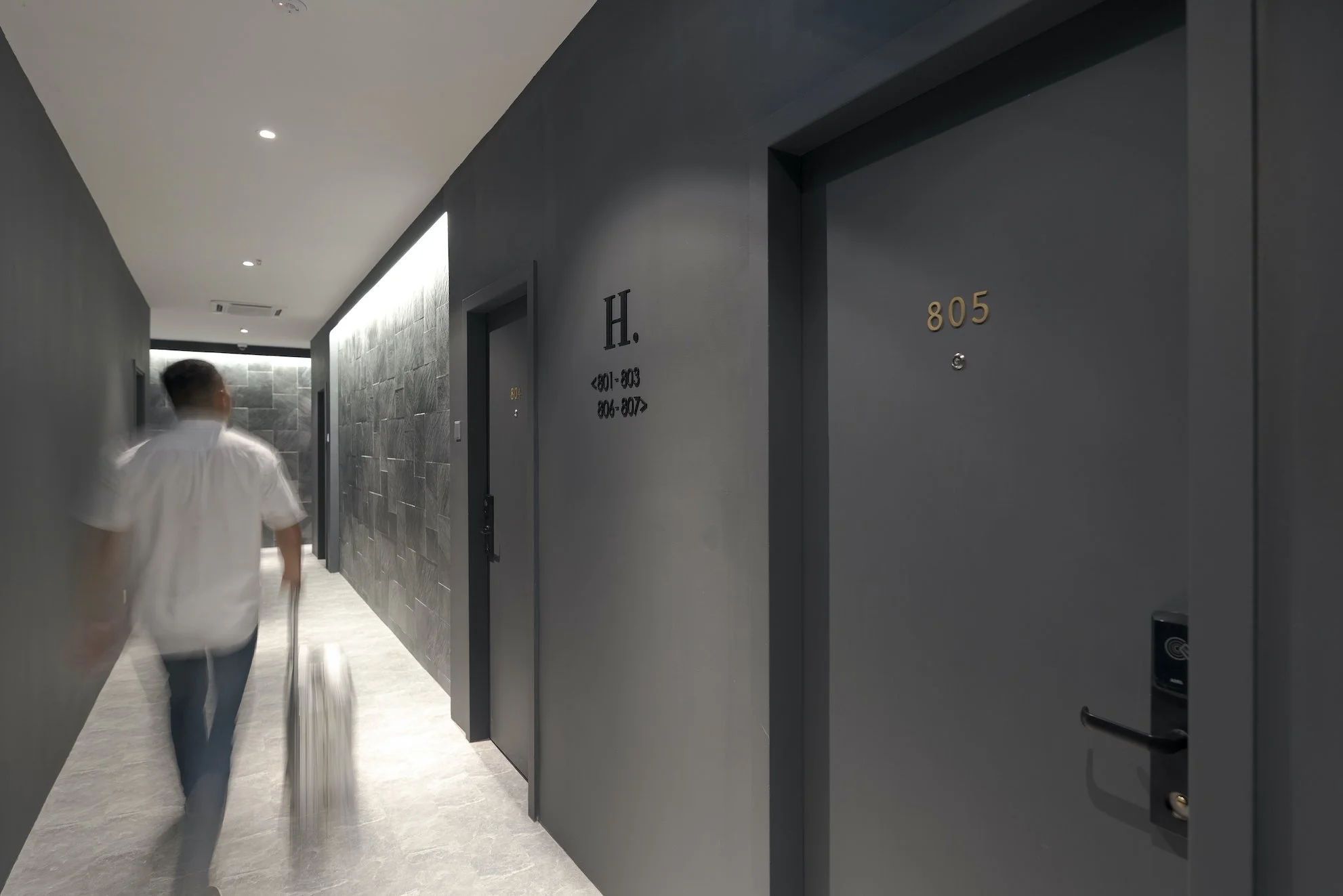 A man walking down a hallway in an apartment building, carrying a suitcase. The hallway has dark gray walls, with room numbers 805 and 806-807 visible on closed doors, and directional signs indicating room ranges.