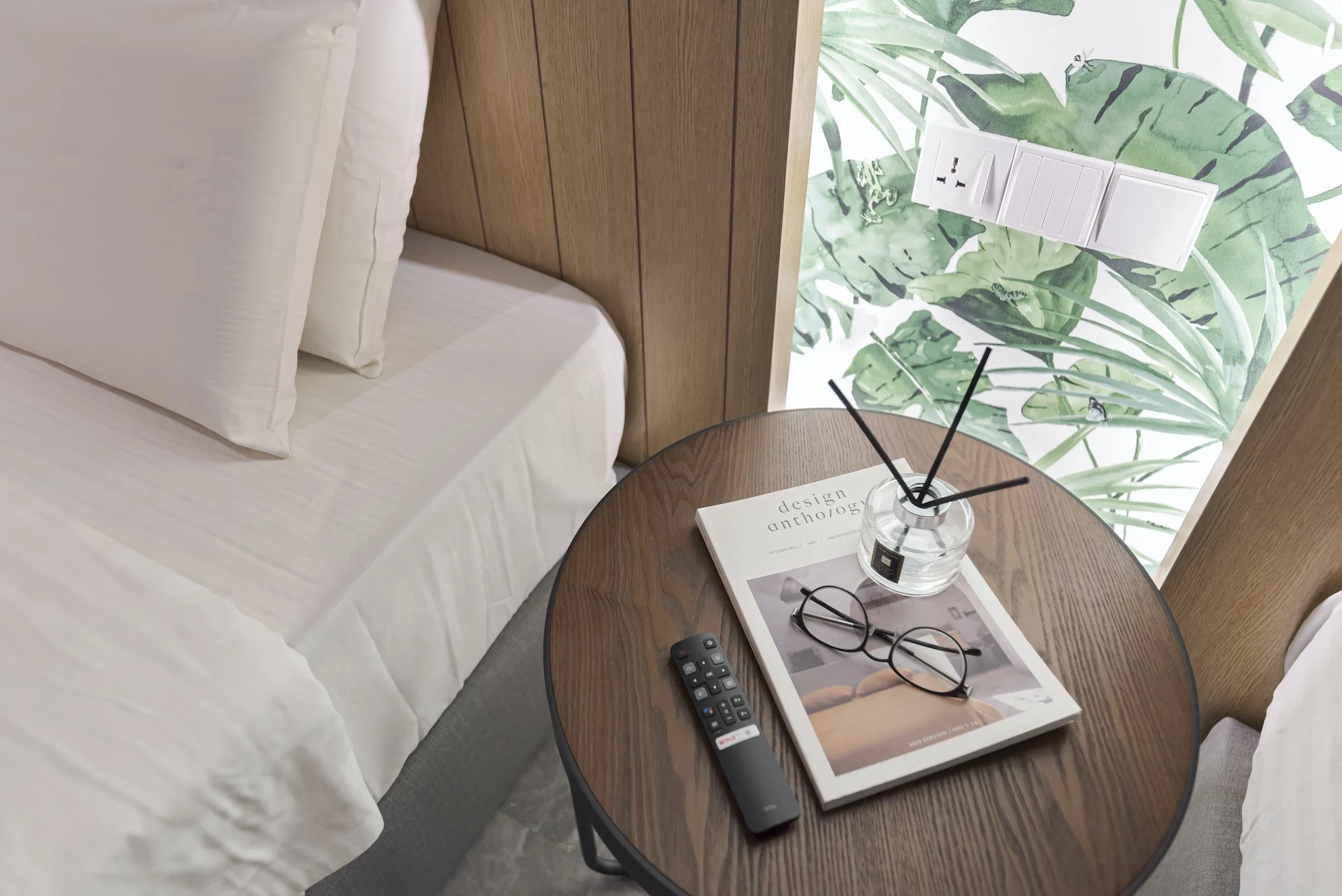 A bedside table between two beds with white bedding, holding a remote control, a magazine, glasses, and a glass vase with reed diffusers. Behind the table, there is a leafy green wallpaper and an electrical outlet with switches.