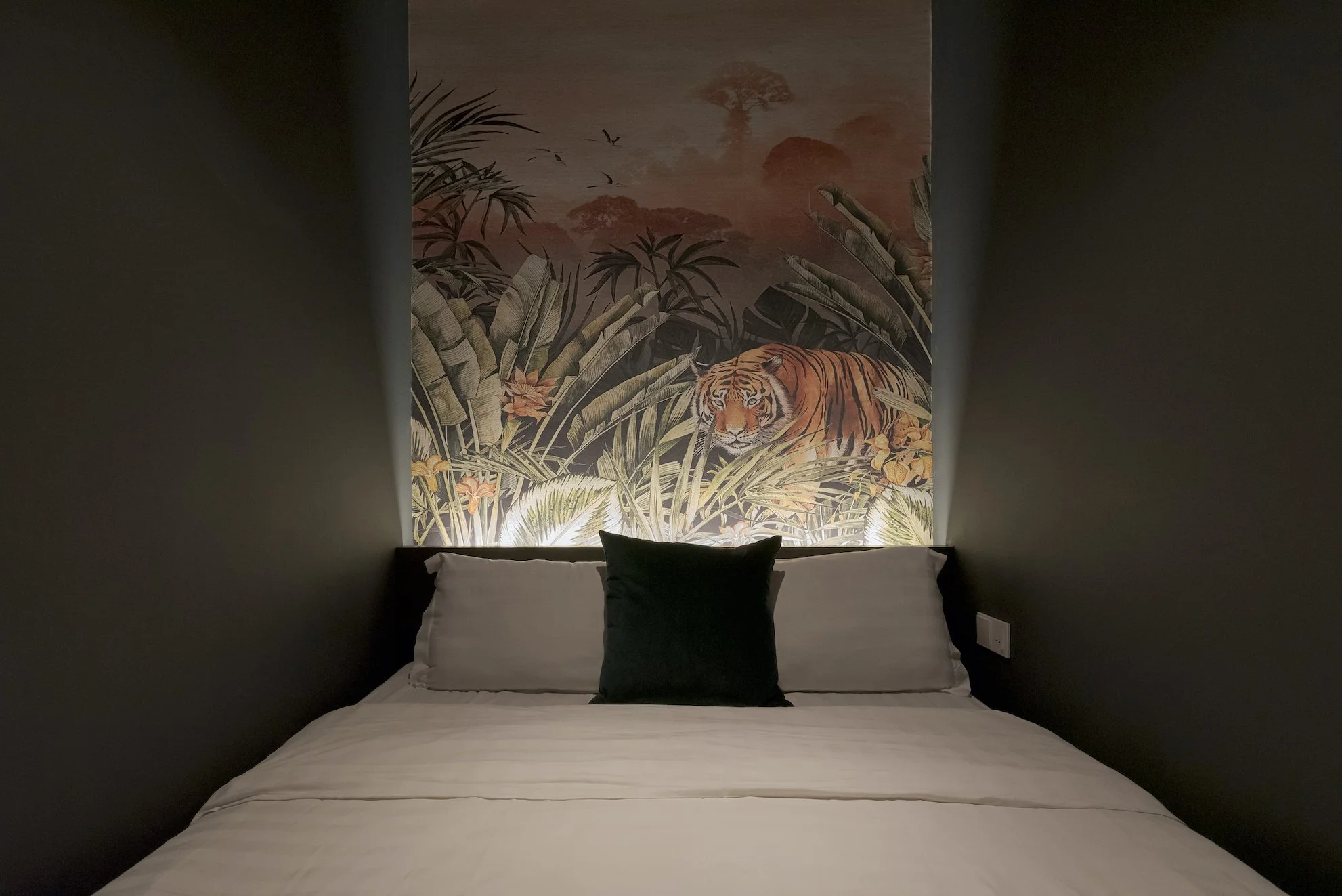 A bed with white sheets and pillows, a black pillow in the center, and a jungle-themed mural of a tiger surrounded by plants on the wall behind.