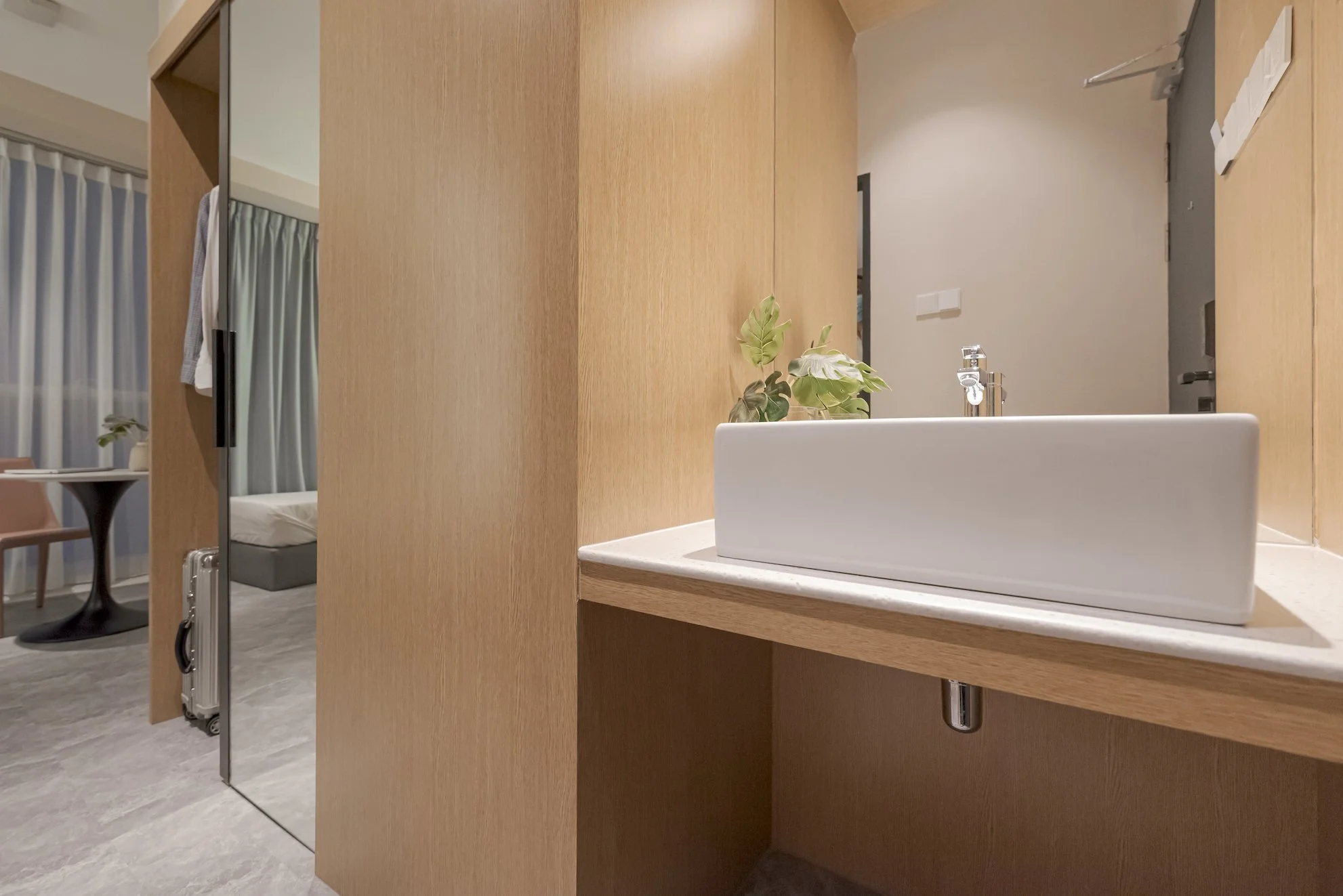 Modern bathroom with wooden vanity, white rectangular sink, and greenery, with room visible in the background.