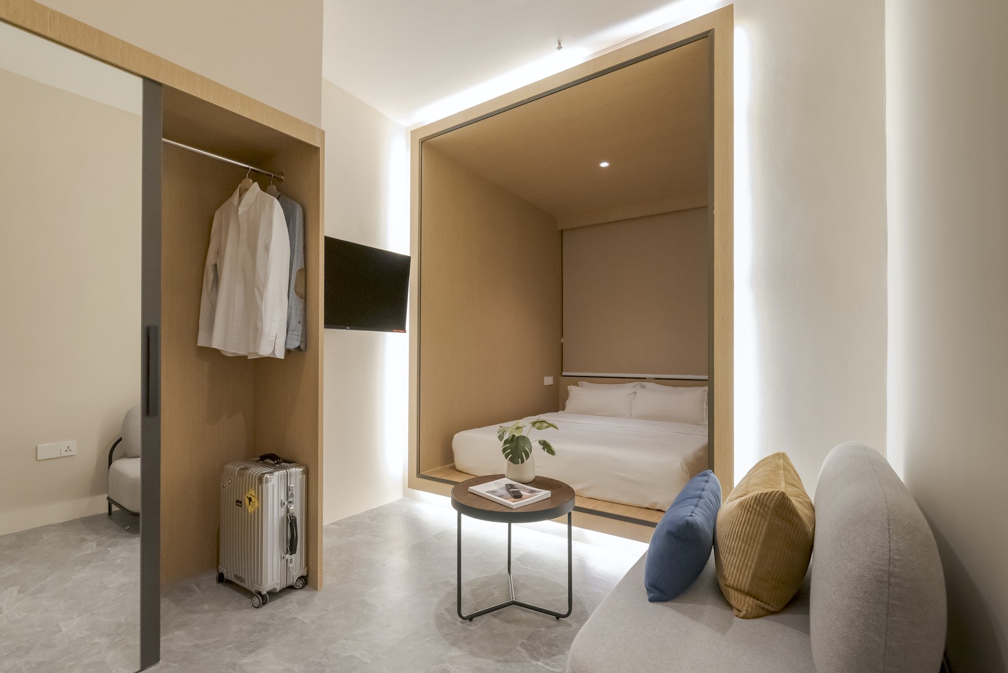 Modern hotel room with a small bed inside a wooden-frame alcove, a gray sofa with colorful pillows, a round side table with a plant and remote, a mounted flat-screen TV, and an open wardrobe with hanging clothes and a suitcase.