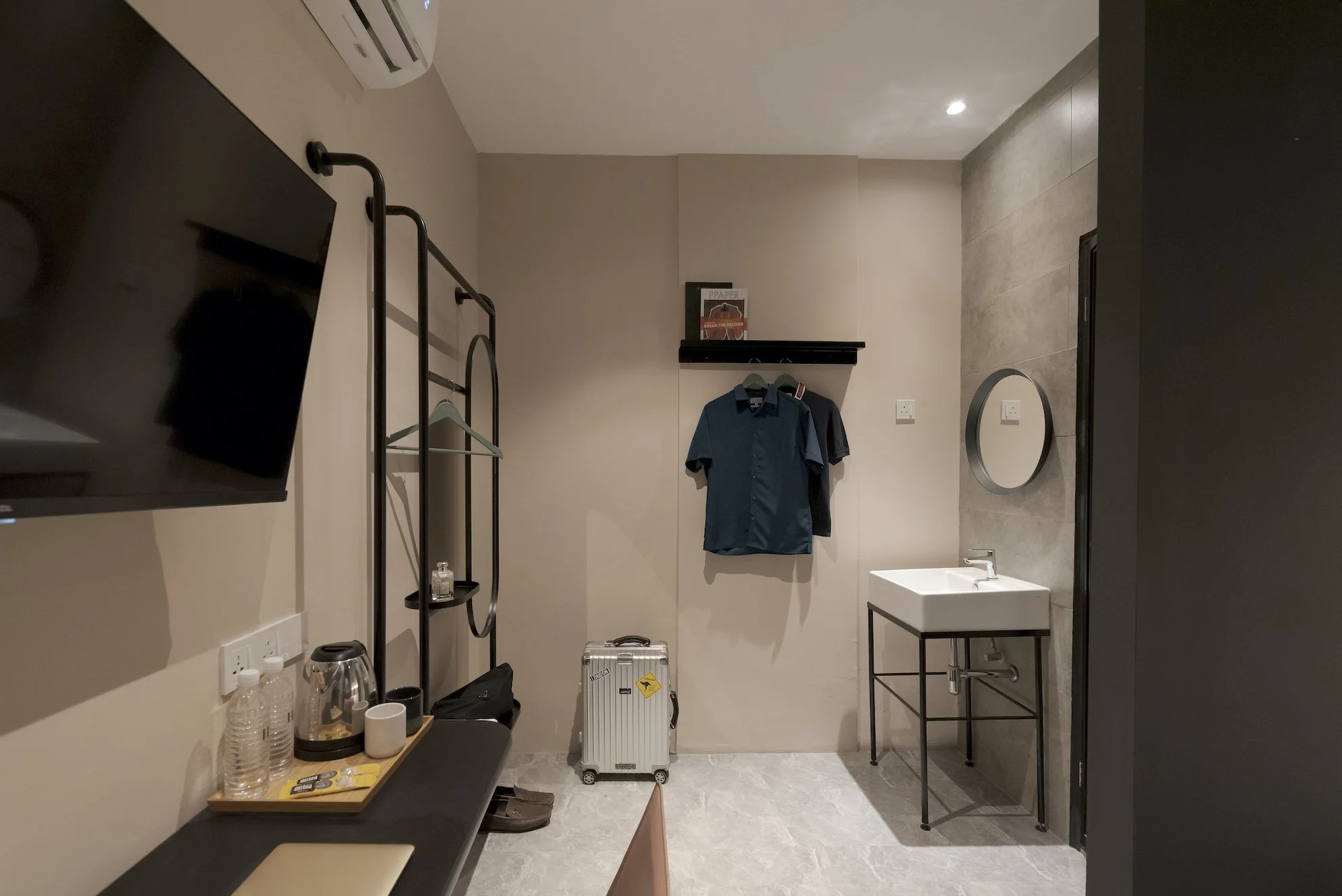 Hotel room with a wall-mounted TV, a metal clothing rack with hanging clothes, a small table with water bottles, a glass of water, a kettle, and cups, a silver suitcase on the floor, a sink with a mirror on the wall, and a black countertop.