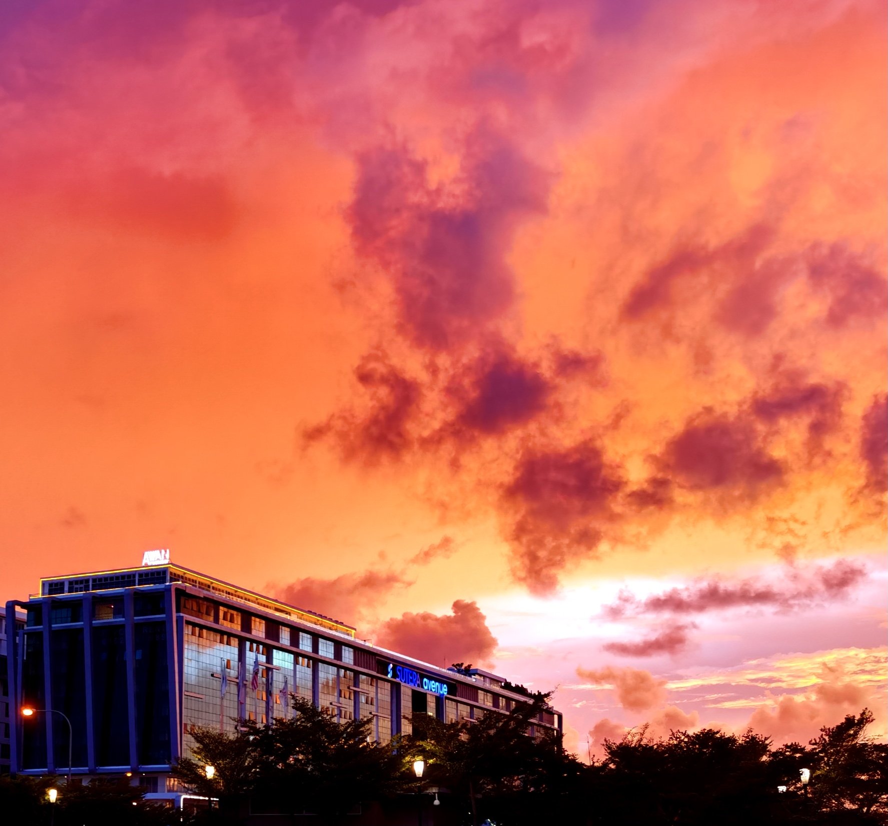 Colorful sunset sky with orange, pink, and purple clouds above a modern building with illuminated signs and trees in the foreground.