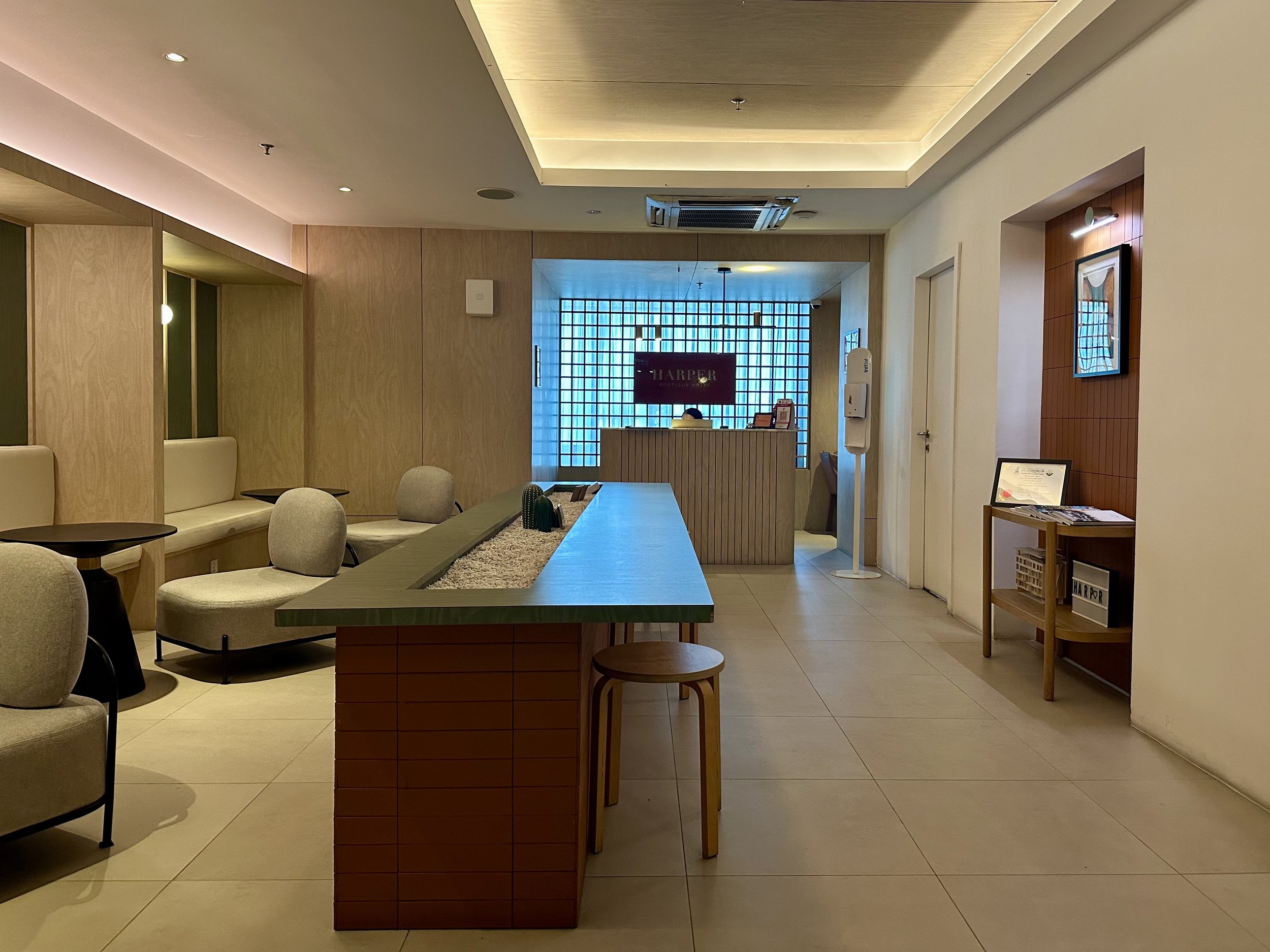 Modern hotel lobby with a reception desk, seating area with beige chairs and small tables, light wood accents, and a backlit blue window in the background.