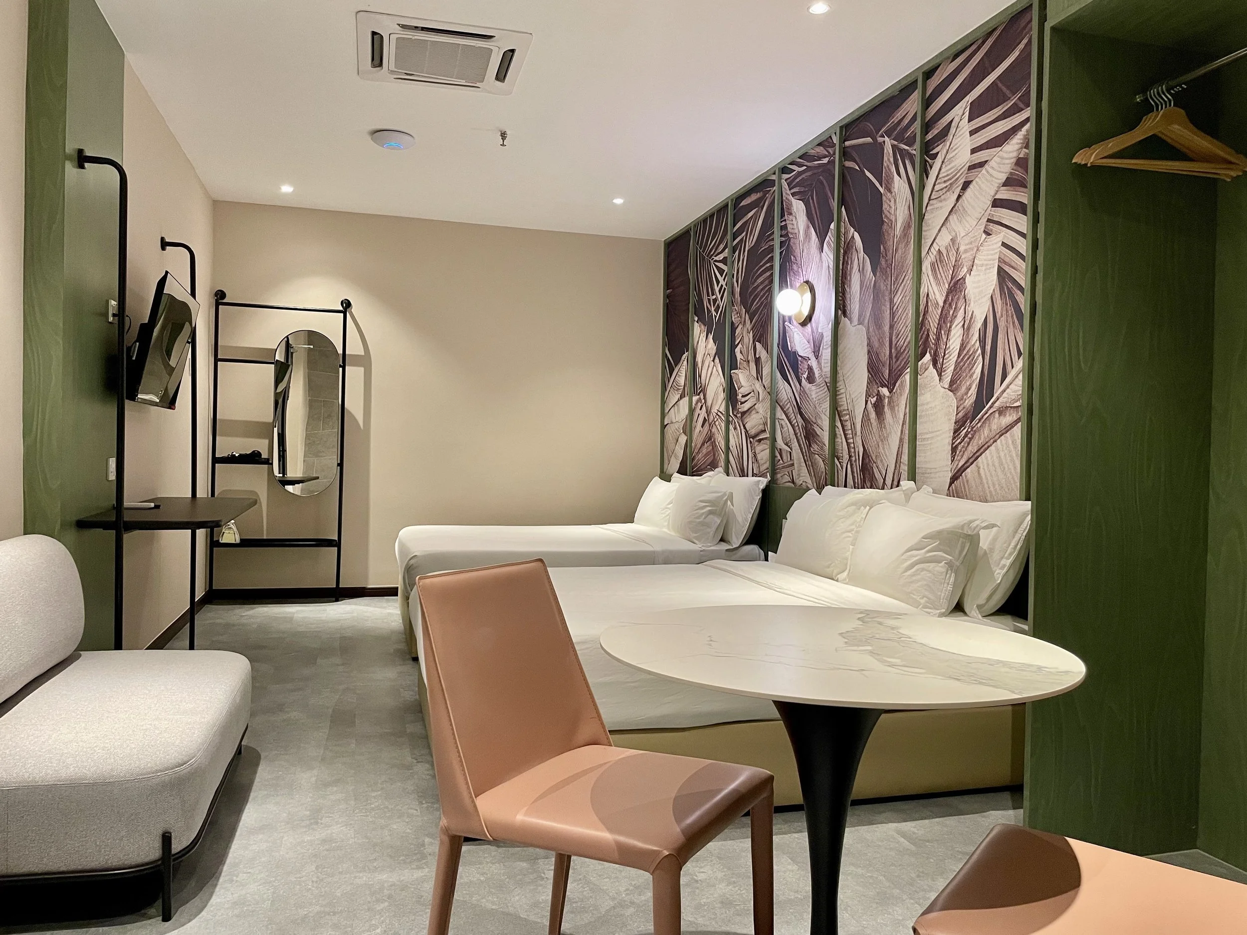 A modern hotel room with two beds, a decorative island theme wall, a small table with pink chairs, a sofa, a mirror, a TV, and closet space. Ceiling air conditioning unit and ceiling lights visible.