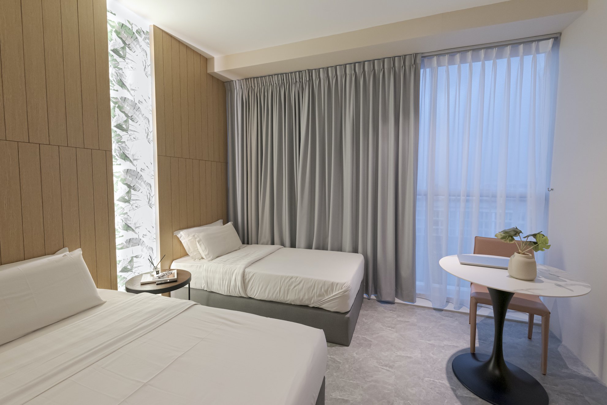 Hotel room with two beds, a small round bedside table with a plant, and a desk with a laptop and a potted plant near large windows with curtains.