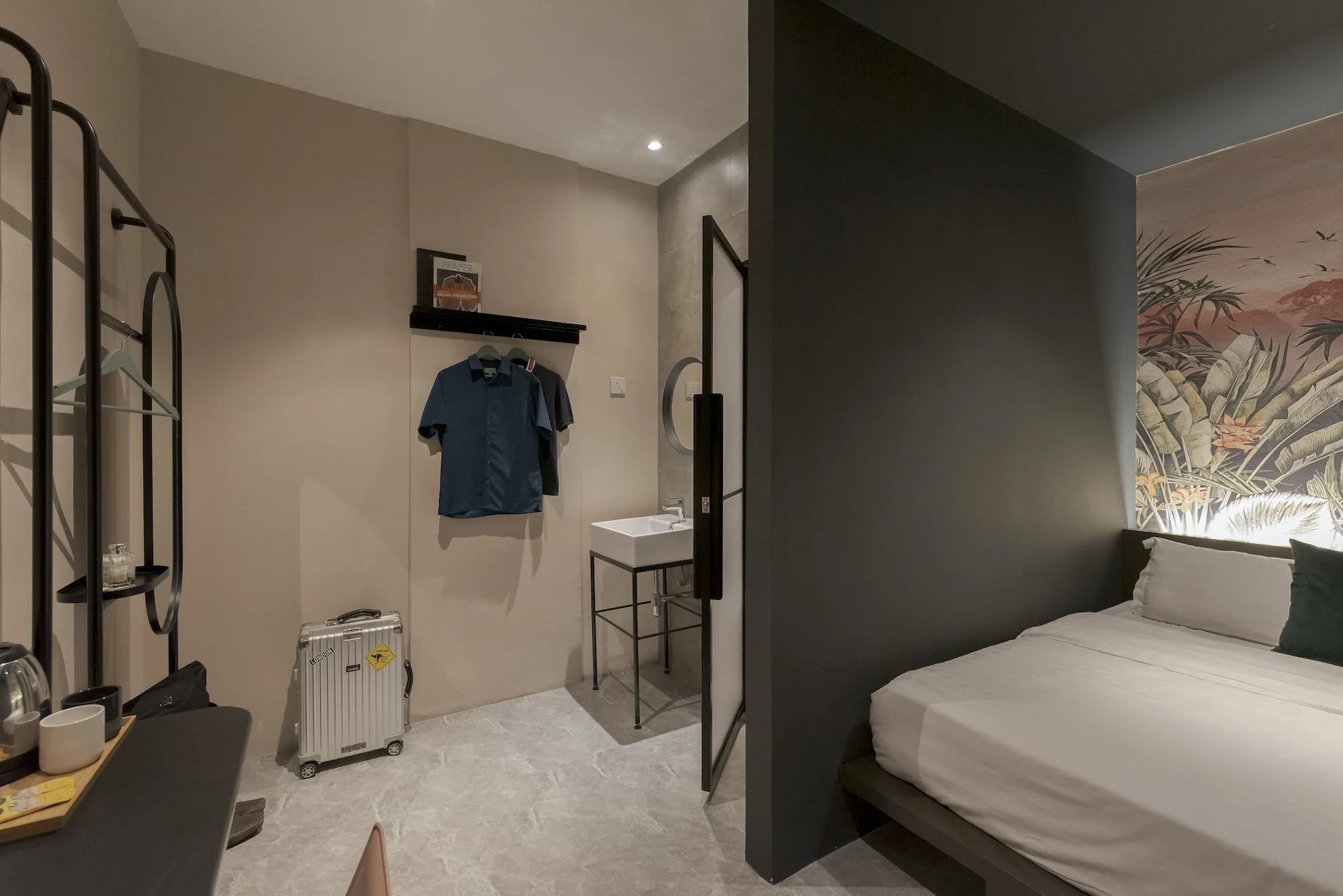 A modern hotel room with a bed, a decorative wall mural, a small sink, a hanging rack with shirts, a suitcase, a table with cups, and a seating area.