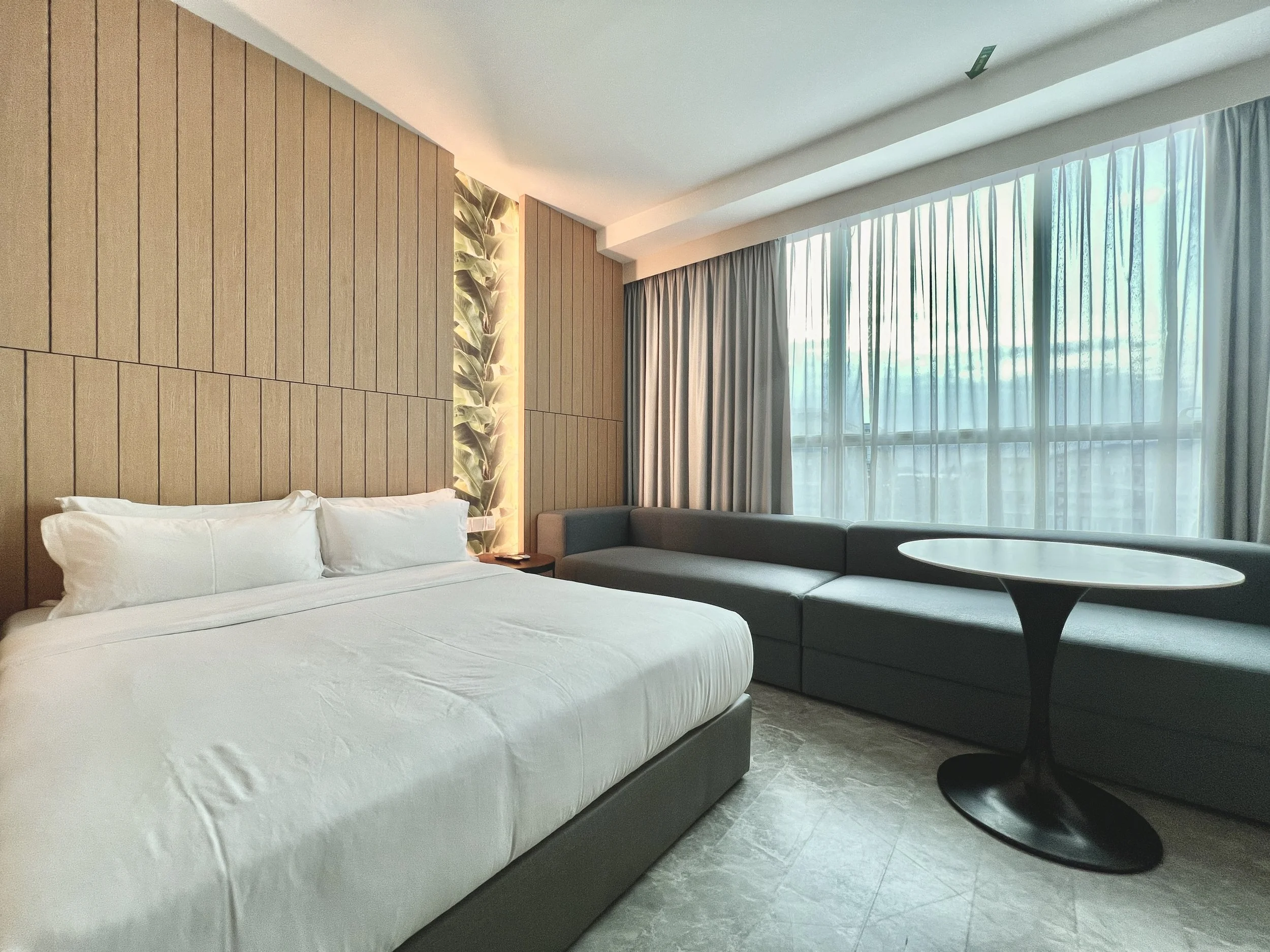 Hotel room with a large bed, wooden accent wall, sofa, round table, and large window with curtains.