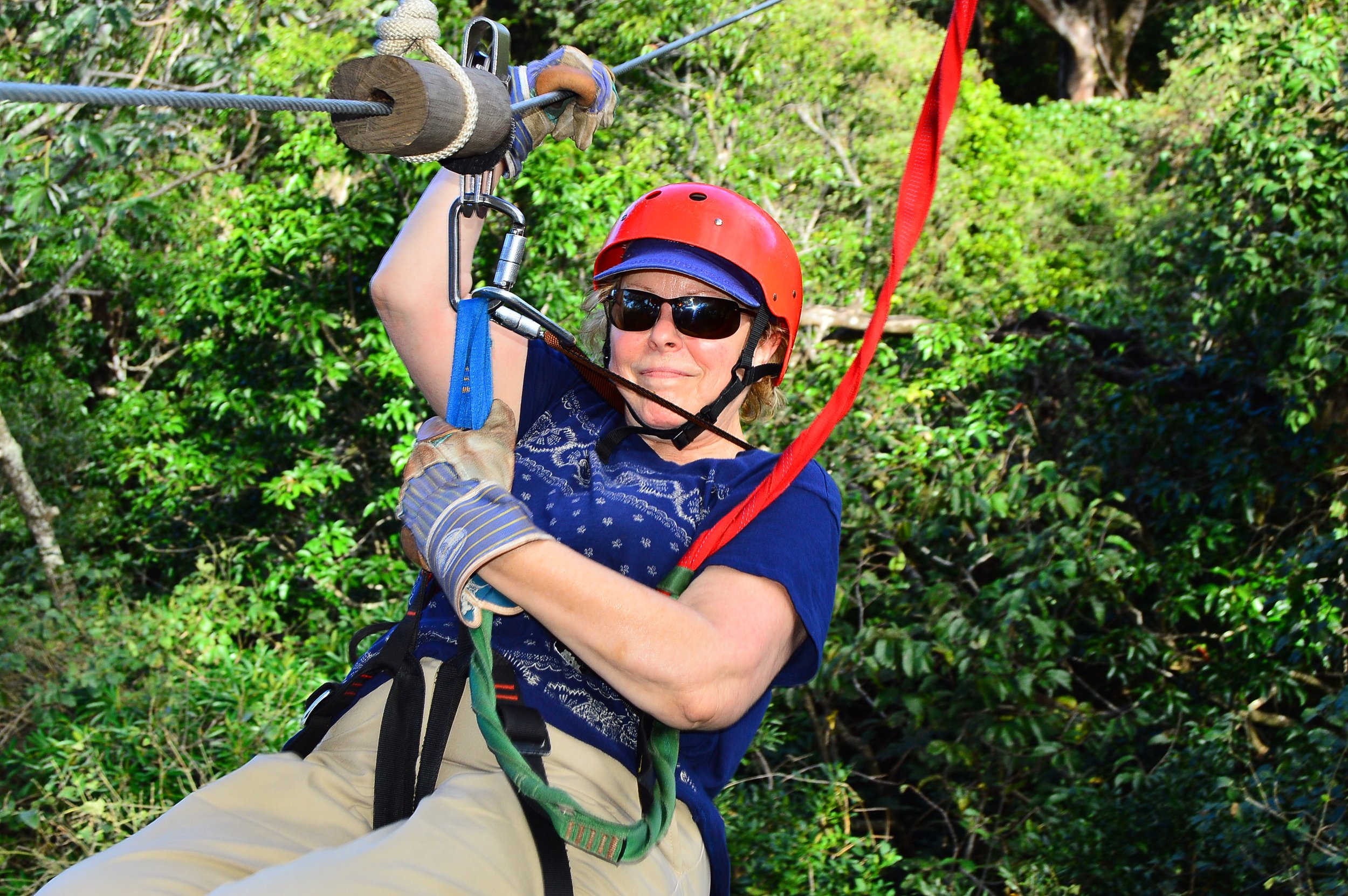 Woman in a red helmet and sunglasses zip lining through a lush green forest.