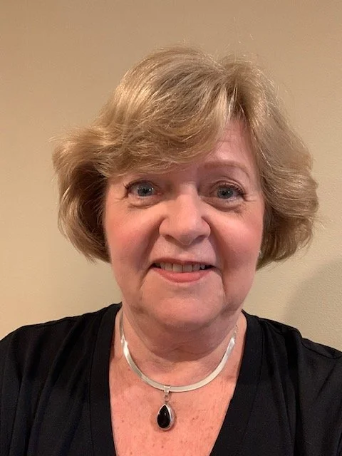 Close-up of a smiling middle-aged woman with short, light brown hair, wearing a black top and a silver necklace with a black pendant.