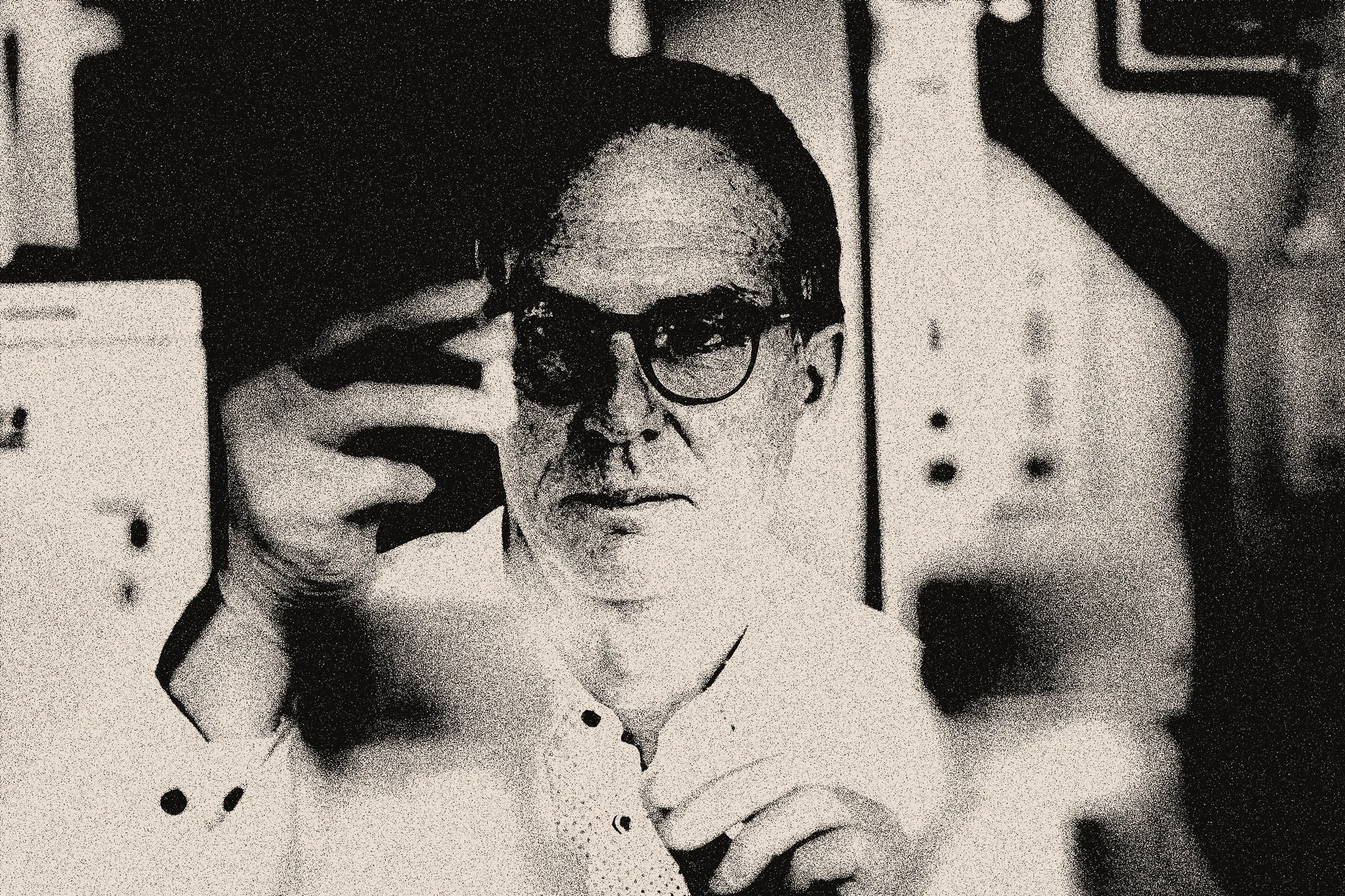 Black and white photo of a man wearing glasses, taking a selfie with a smartphone. He is in an indoor setting with visible modern decor.