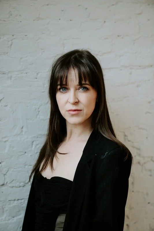A woman with shoulder-length brown hair and bangs standing against a white brick wall, wearing a black blazer and top.