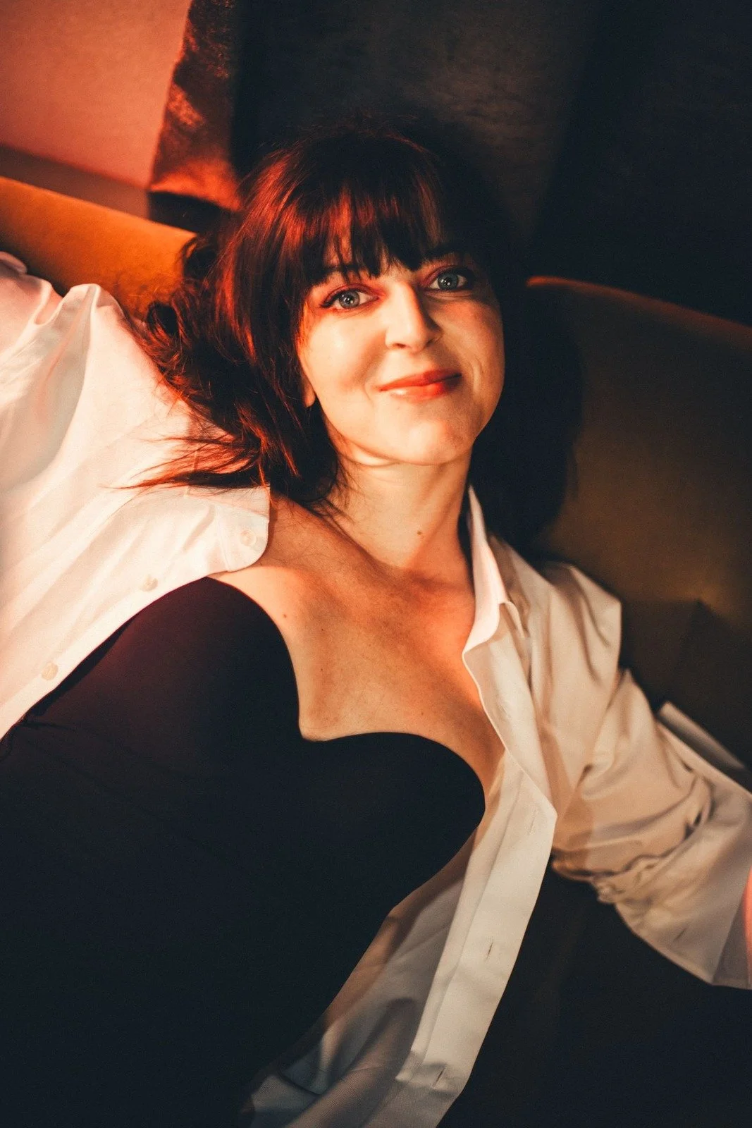 A woman with dark hair and bangs lying on a bed, smiling, wearing a white shirt and a black off-shoulder top.