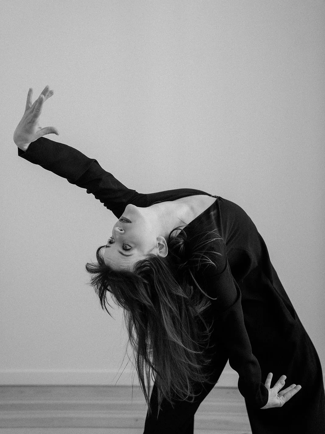 A woman with long hair in a black outfit is leaning backward with her left arm extended upward and her right arm touching the ground, striking a dance or yoga pose against a plain background.