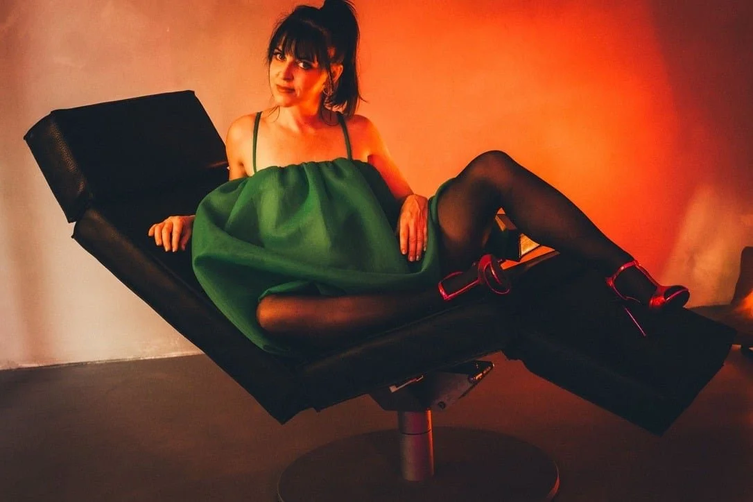 Woman in a green dress and black stockings reclining on a black chair against a red-lit background.