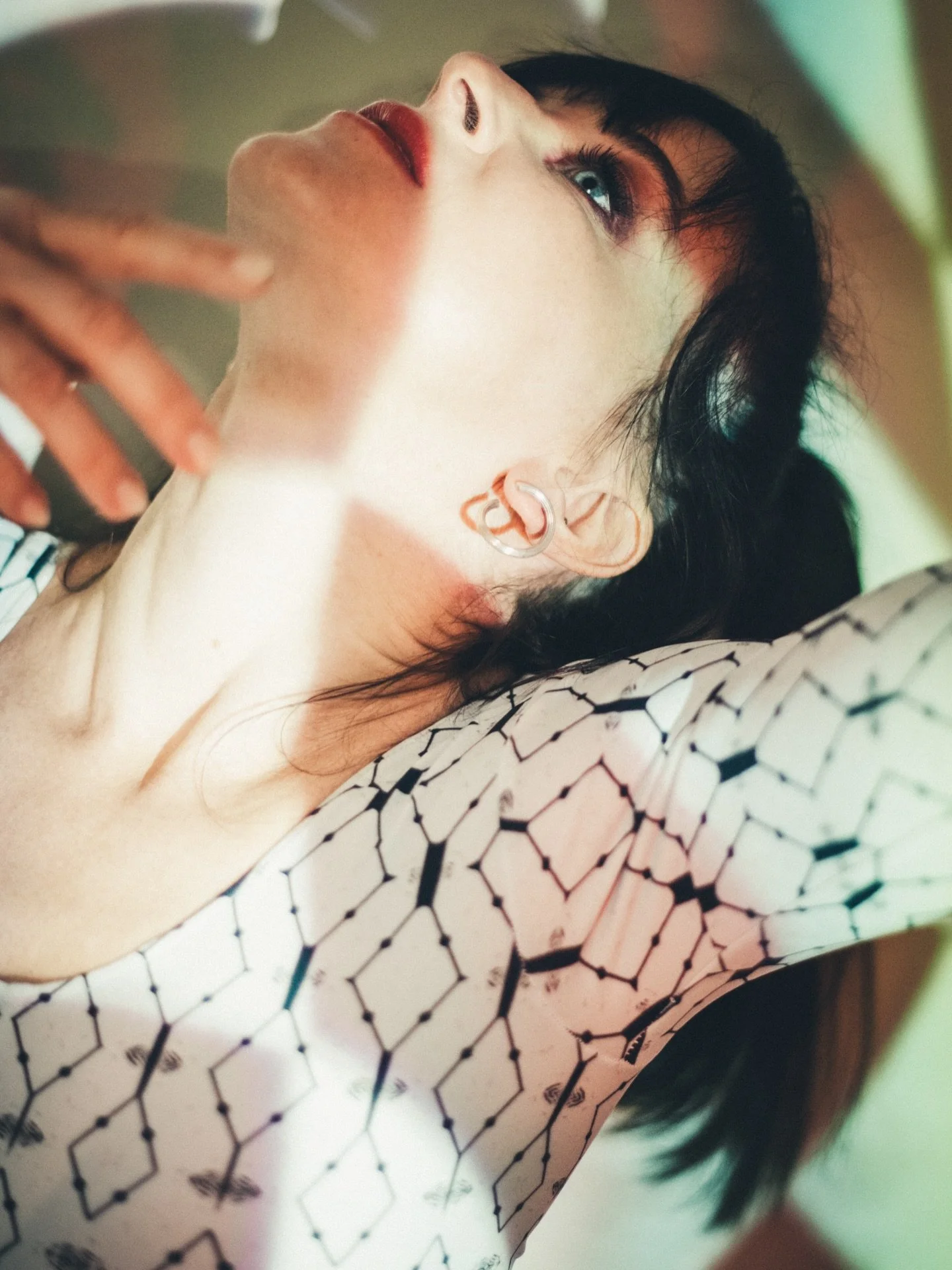 Close-up of a woman with dark hair and blue eyes, wearing earrings and a black and white patterned top, lying on a surface.
