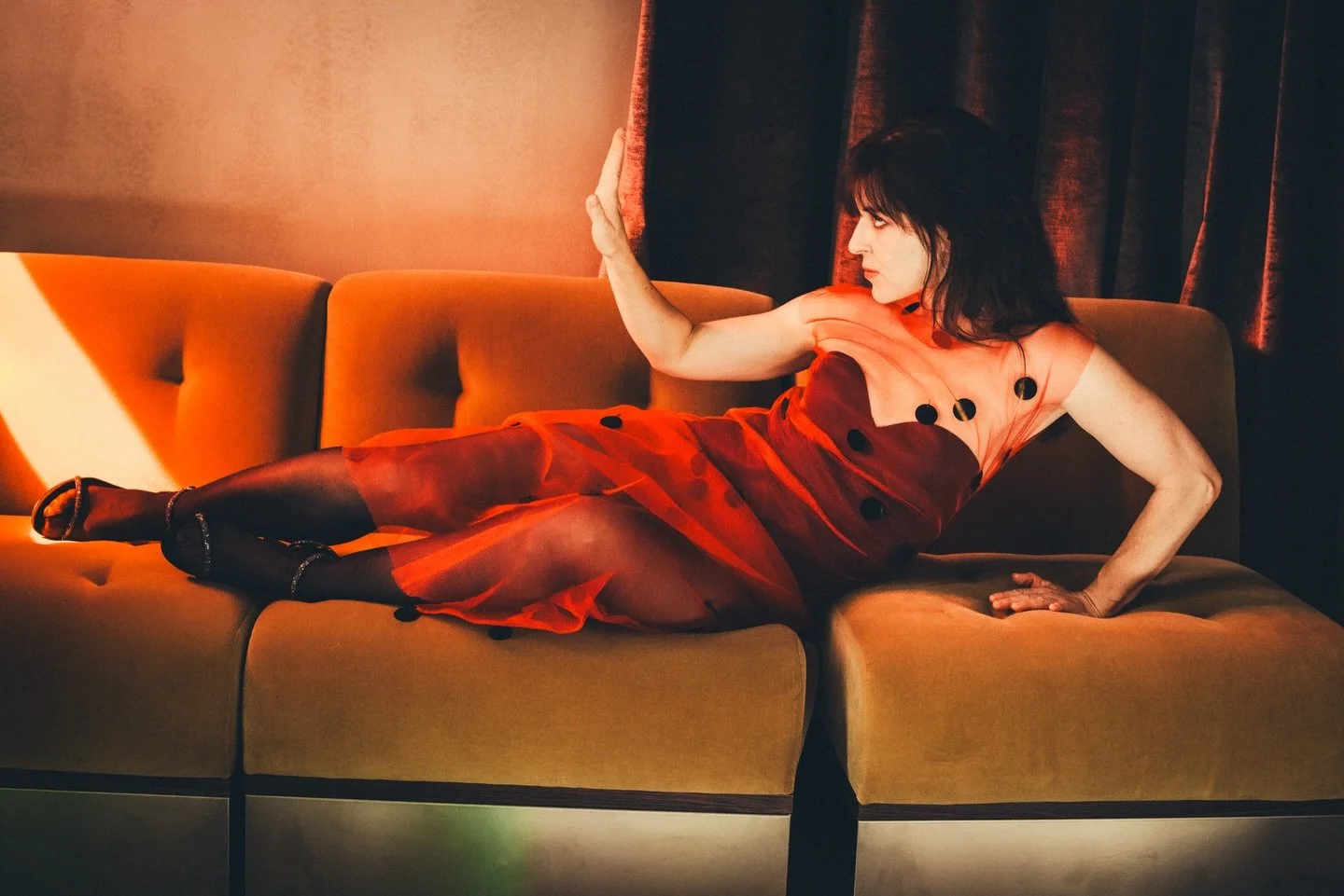 A woman in a red dress with black polka dots is reclining on an orange couch, with her left arm extended and her hand pressed against a dark curtain, looking to her right.