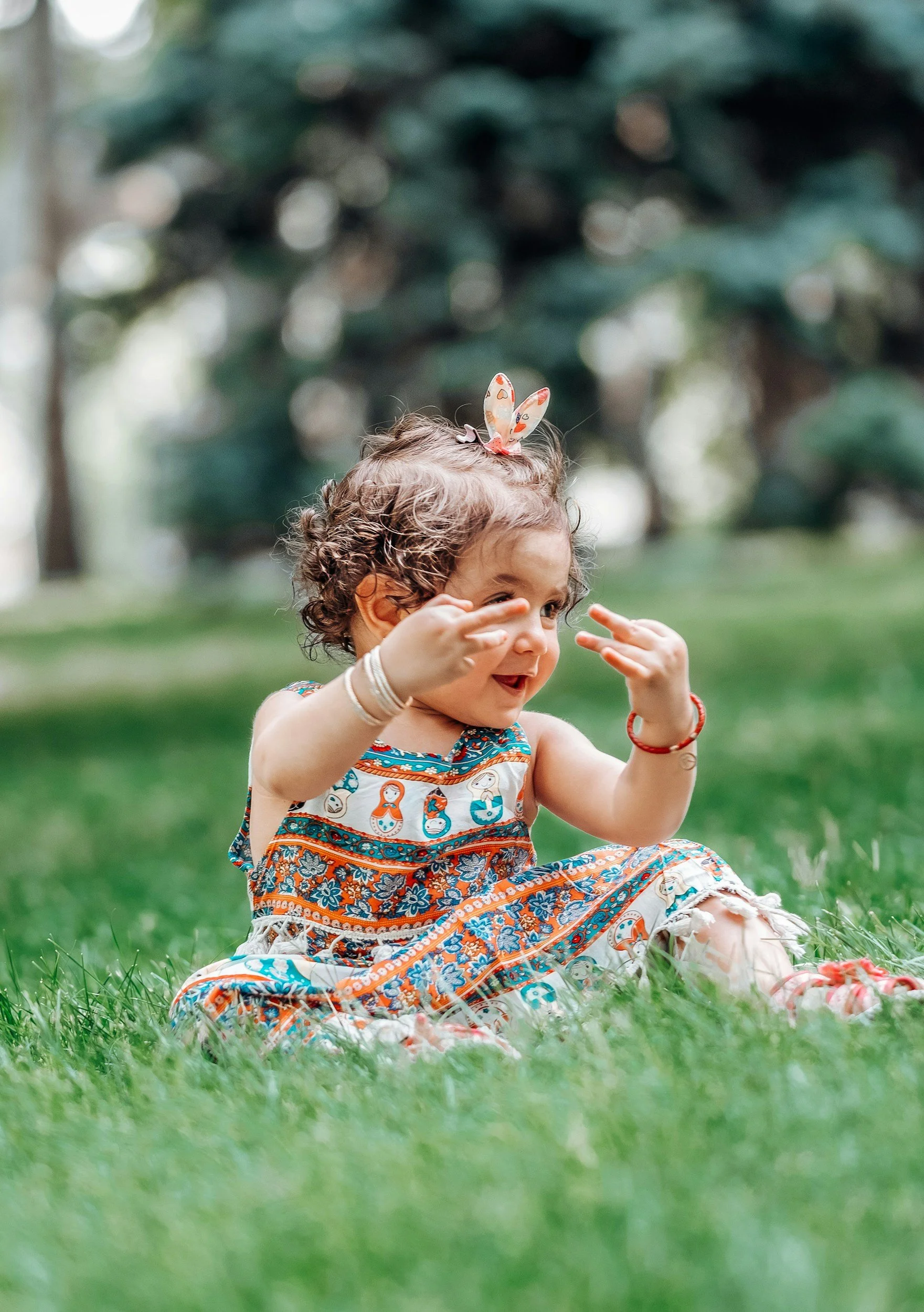 A young girl sitting on green grass outdoors, wearing a colorful dress with a Matryoshka doll pattern, and a hair bow. She is smiling and playing with her hands, with a blurred background of trees.