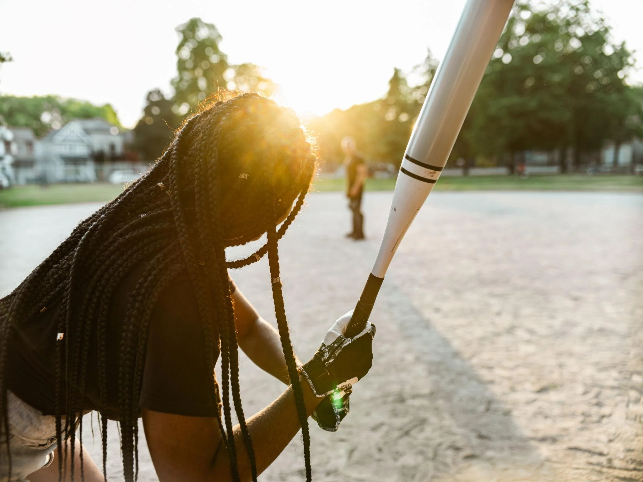A woman with braids at a baseball field holding a baseball bat, with the sun setting in the background.
