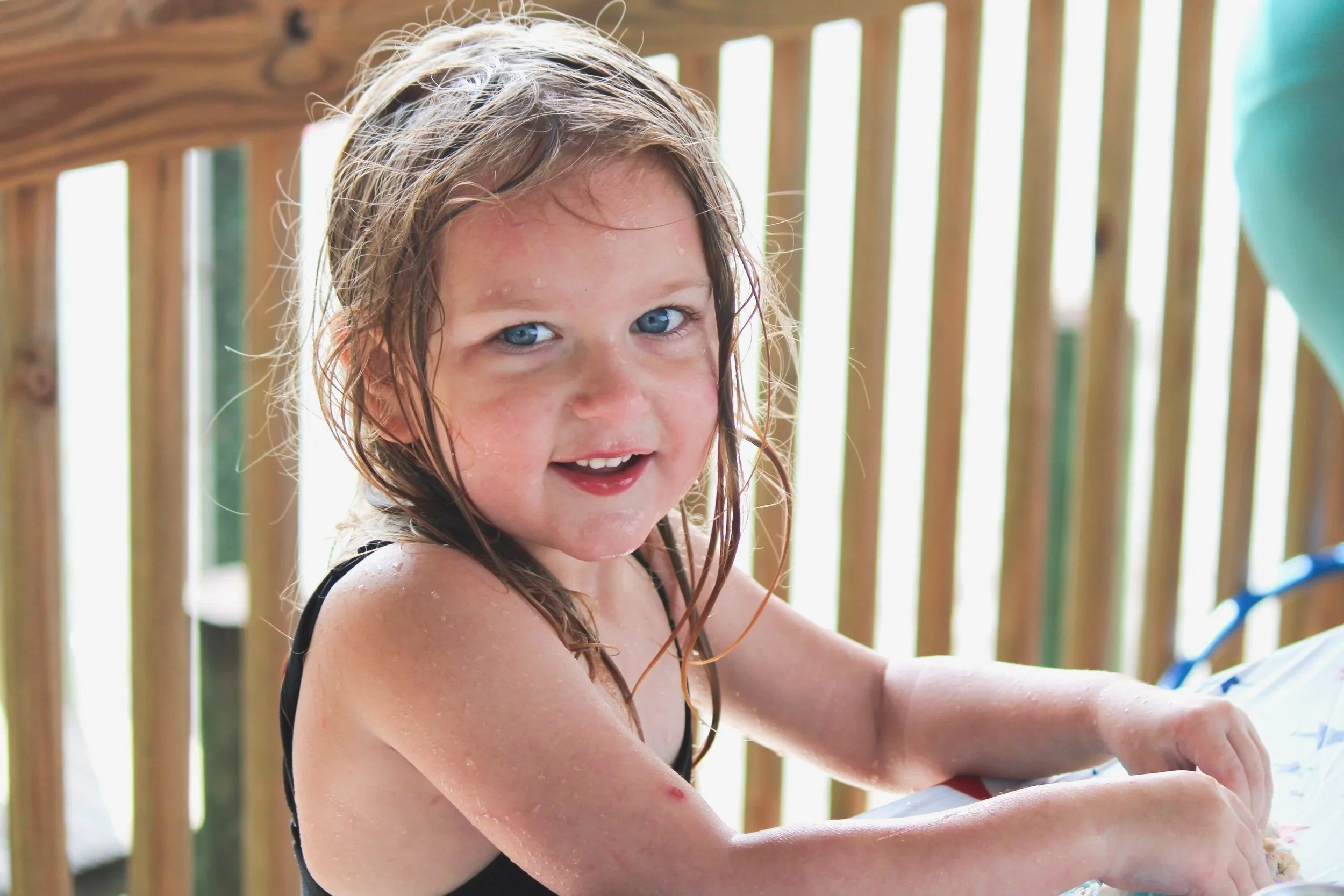 A young girl with wet hair and blue eyes smiling on a wooden deck, wearing a black swimsuit.
