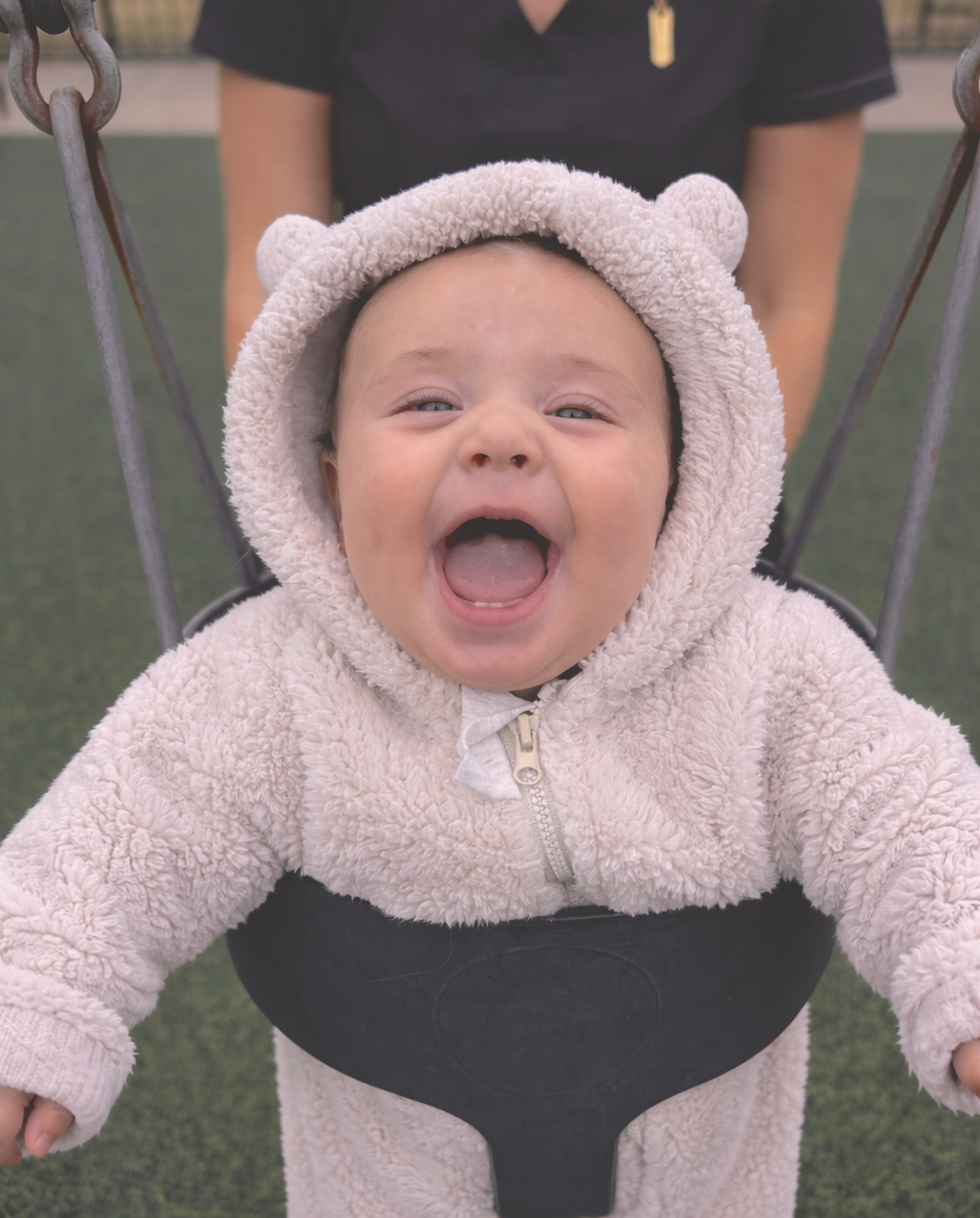 A young child in a soft, fluffy bear hoodie smiling and laughing in a swing at a playground.