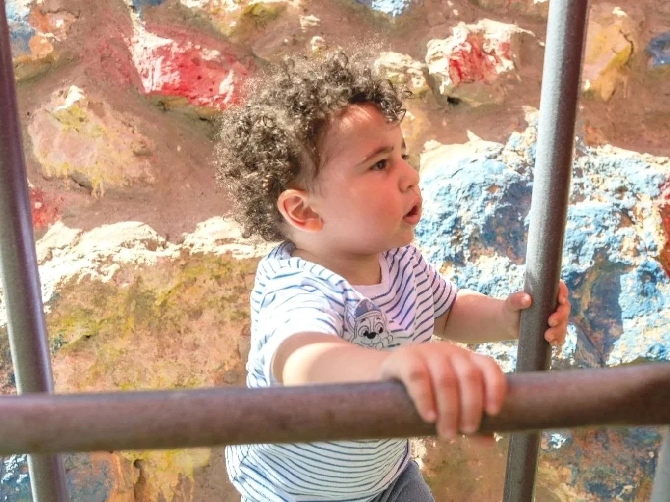 A young child with curly hair climbing on a metal structure at a playground, with a colorful rock wall behind him.