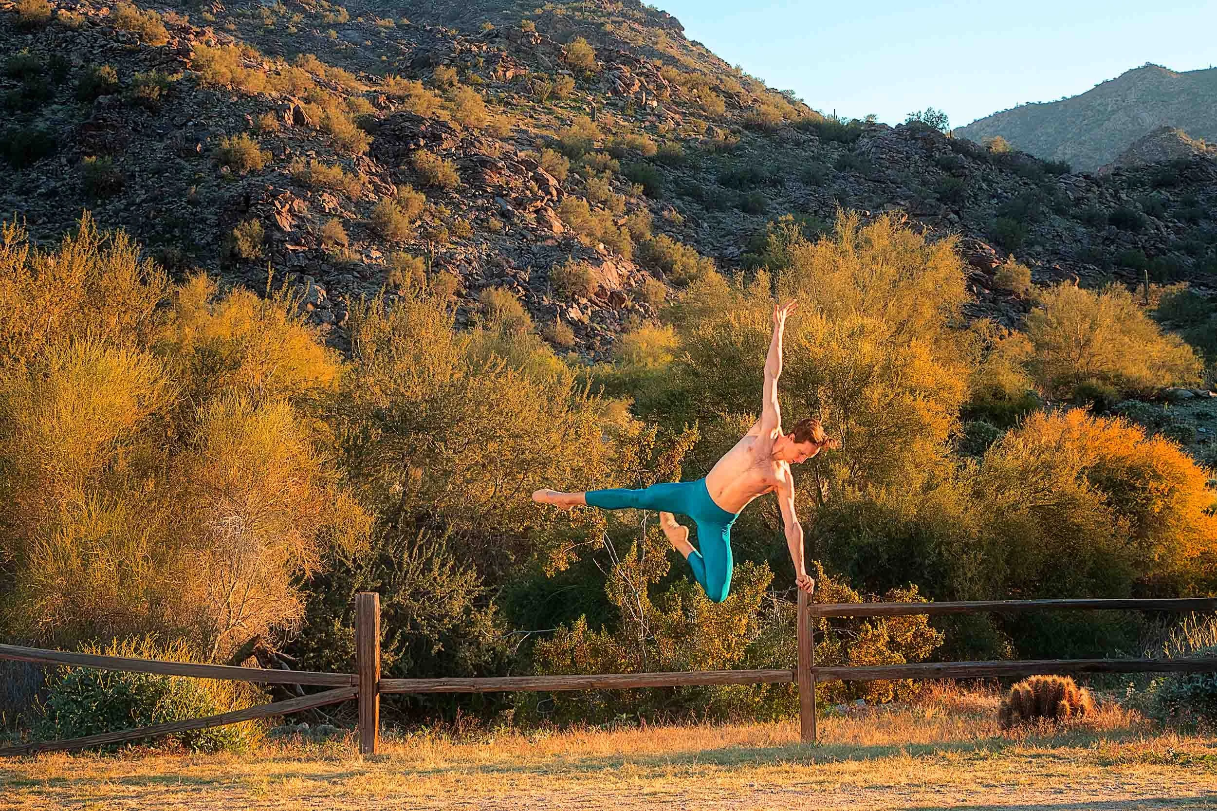 A man in blue gym wear performing a handstand on a trampoline in a desert landscape with mountains and trees during sunset.
