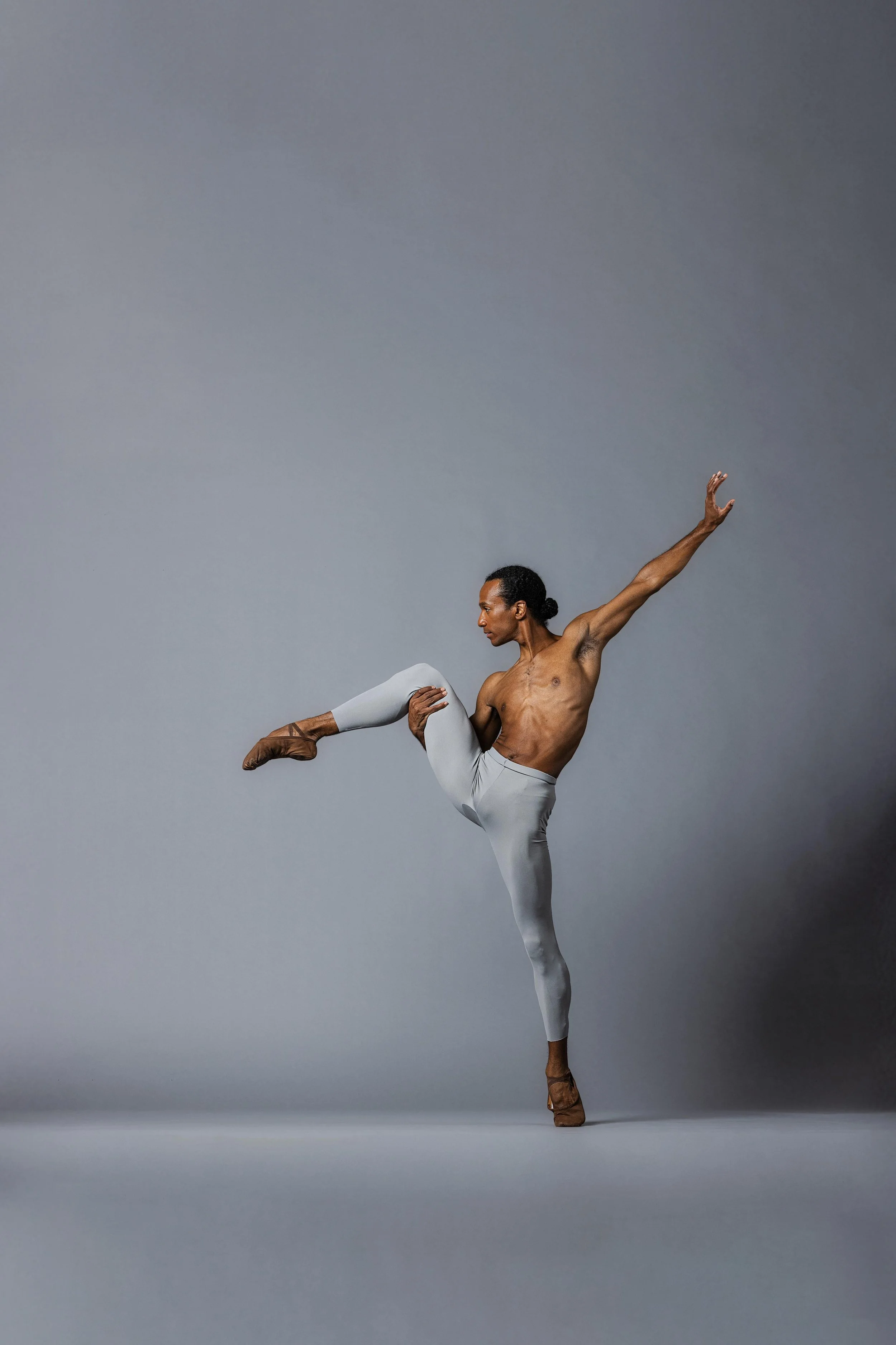 A male ballet dancer performing a ballet pose against a grey background, shirtless and wearing light grey tights with brown ballet slippers.
