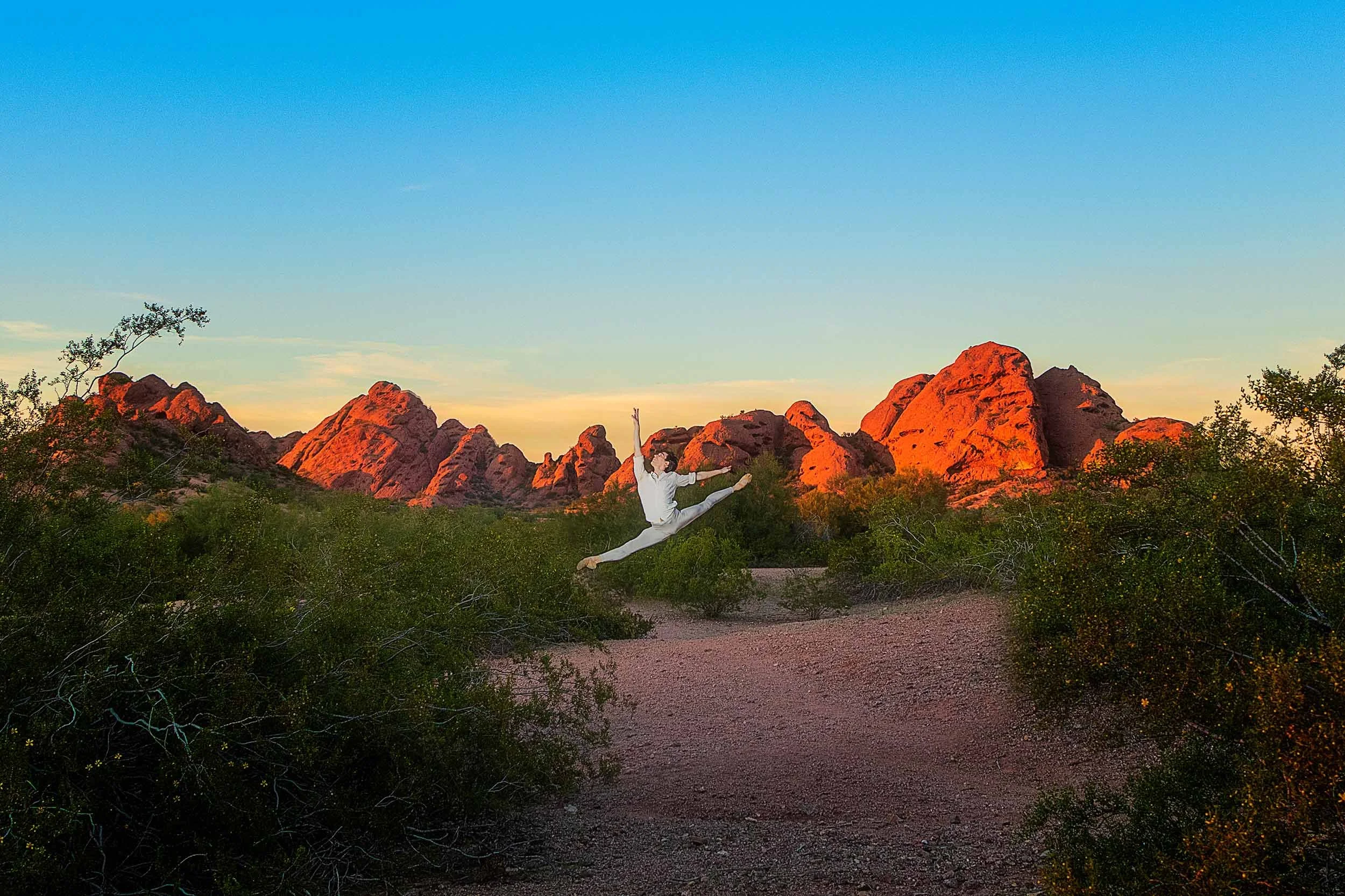 A woman performing a leap in a desert landscape with red rocks and green bushes during sunset or sunrise