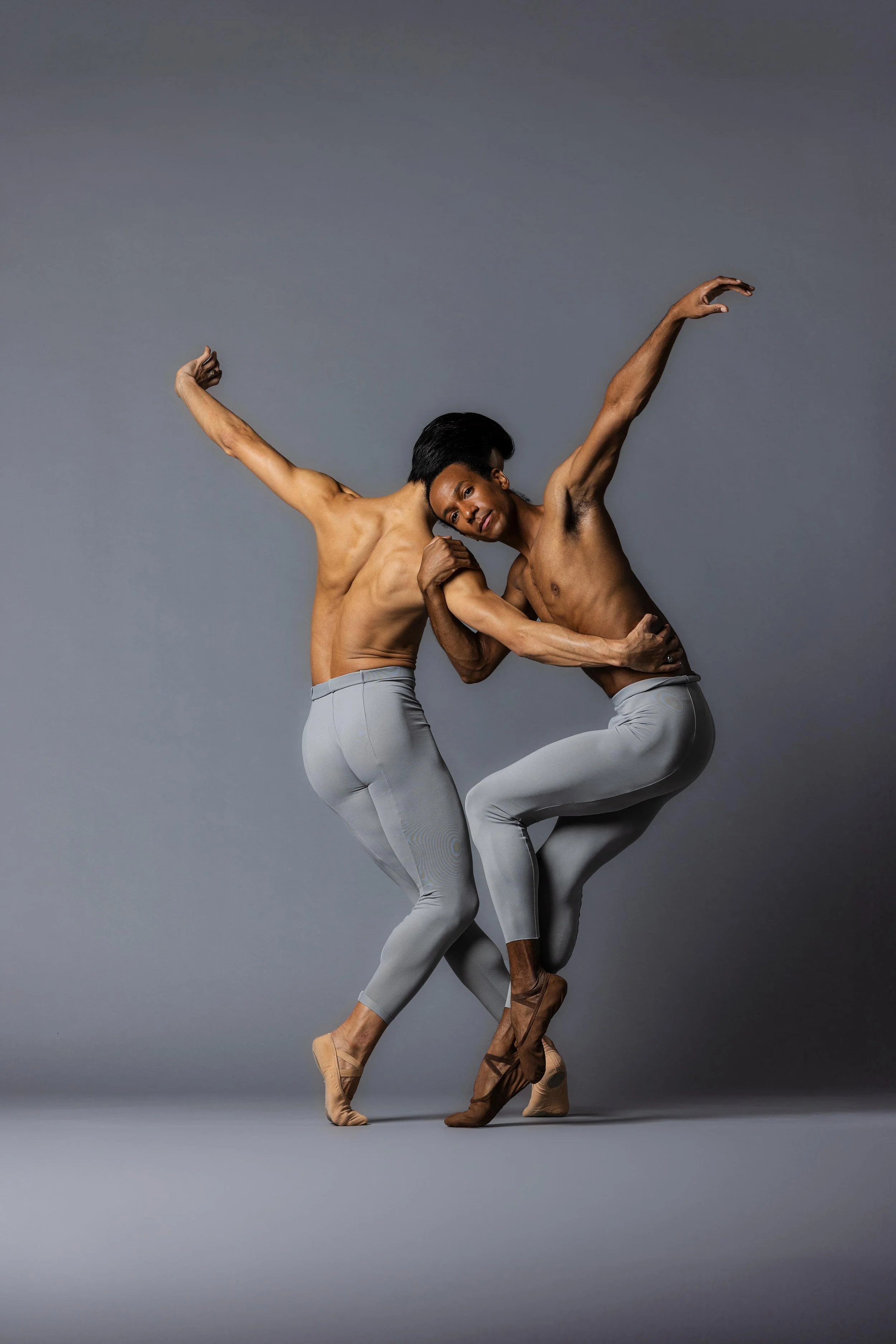 Two male ballet dancers perform a dance pose against a gray background.