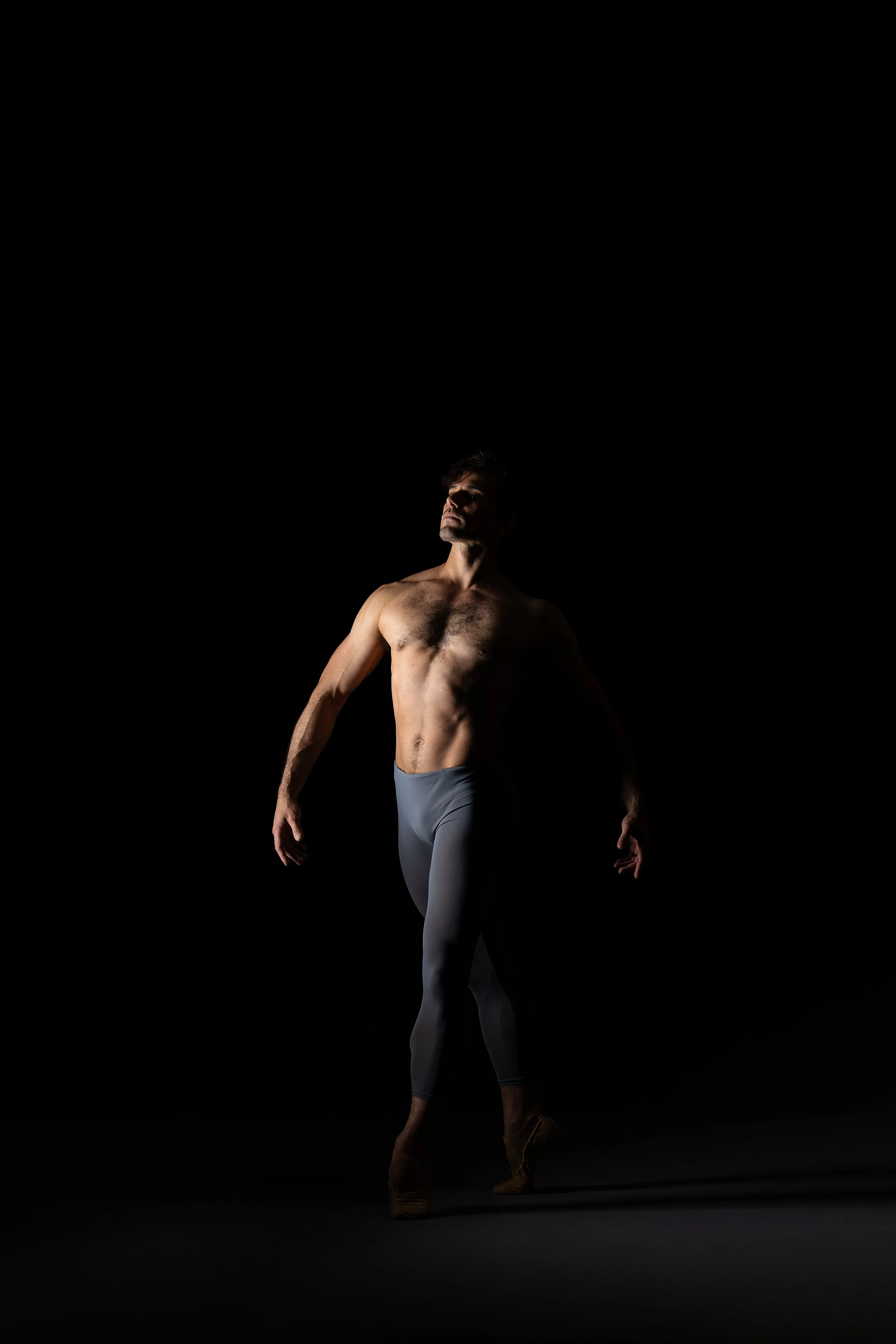 A male ballet dancer with a muscular physique posing shirtless against a dark background, wearing gray tights and dance shoes, illuminated by dramatic lighting.