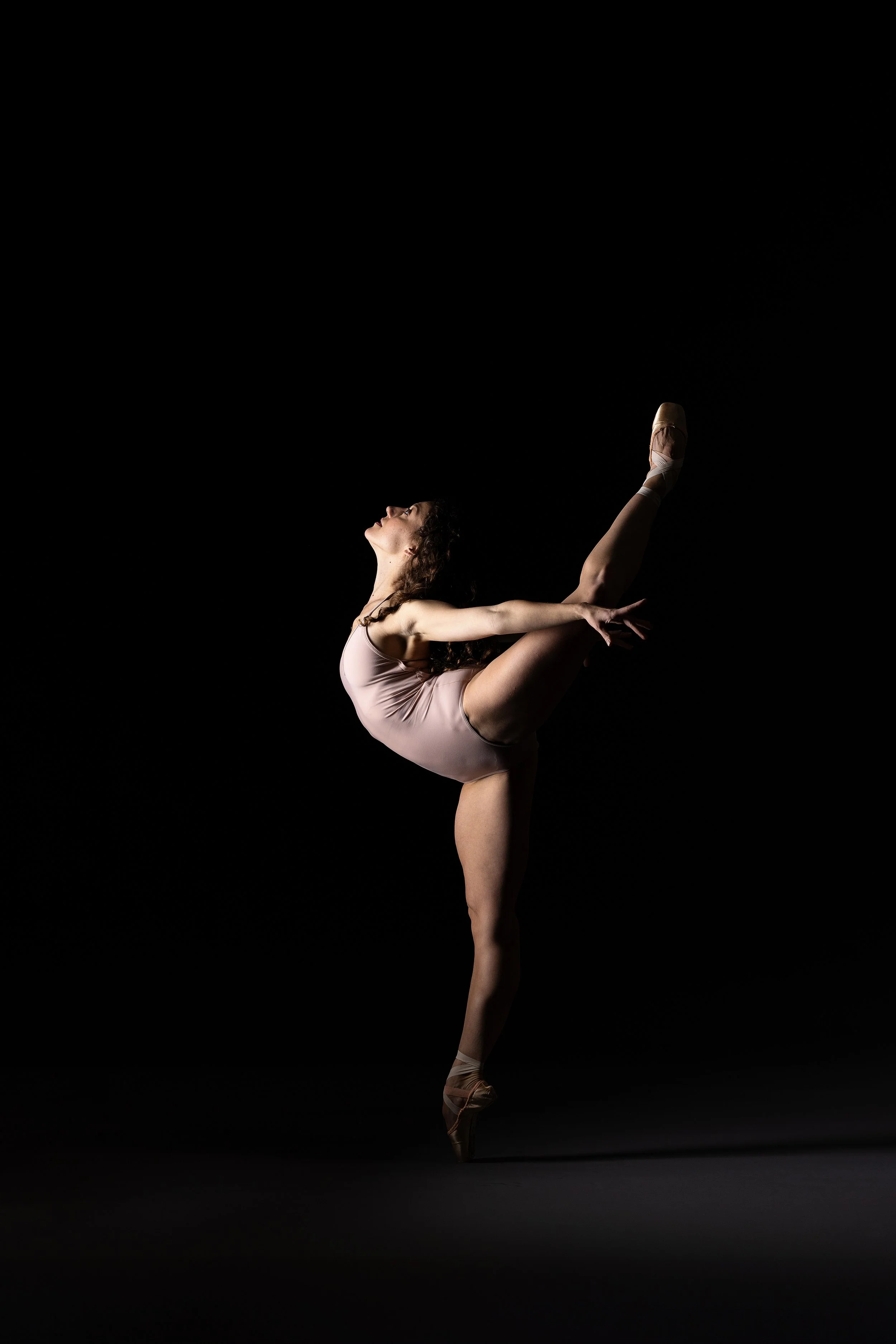 Ballet dancer performing an arabesque pose in a pink leotard against a black background.