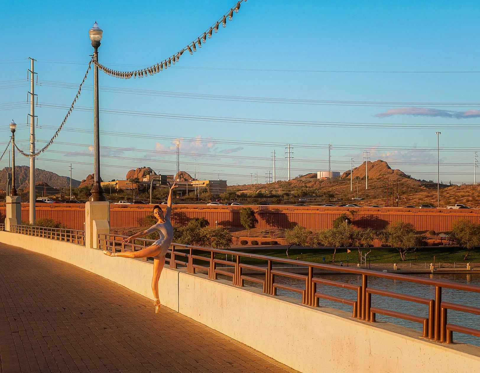 A ballet dancer in a gray dress performs a dance pose on a paved walkway near a river, with a cityscape and mountains in the background during sunset.