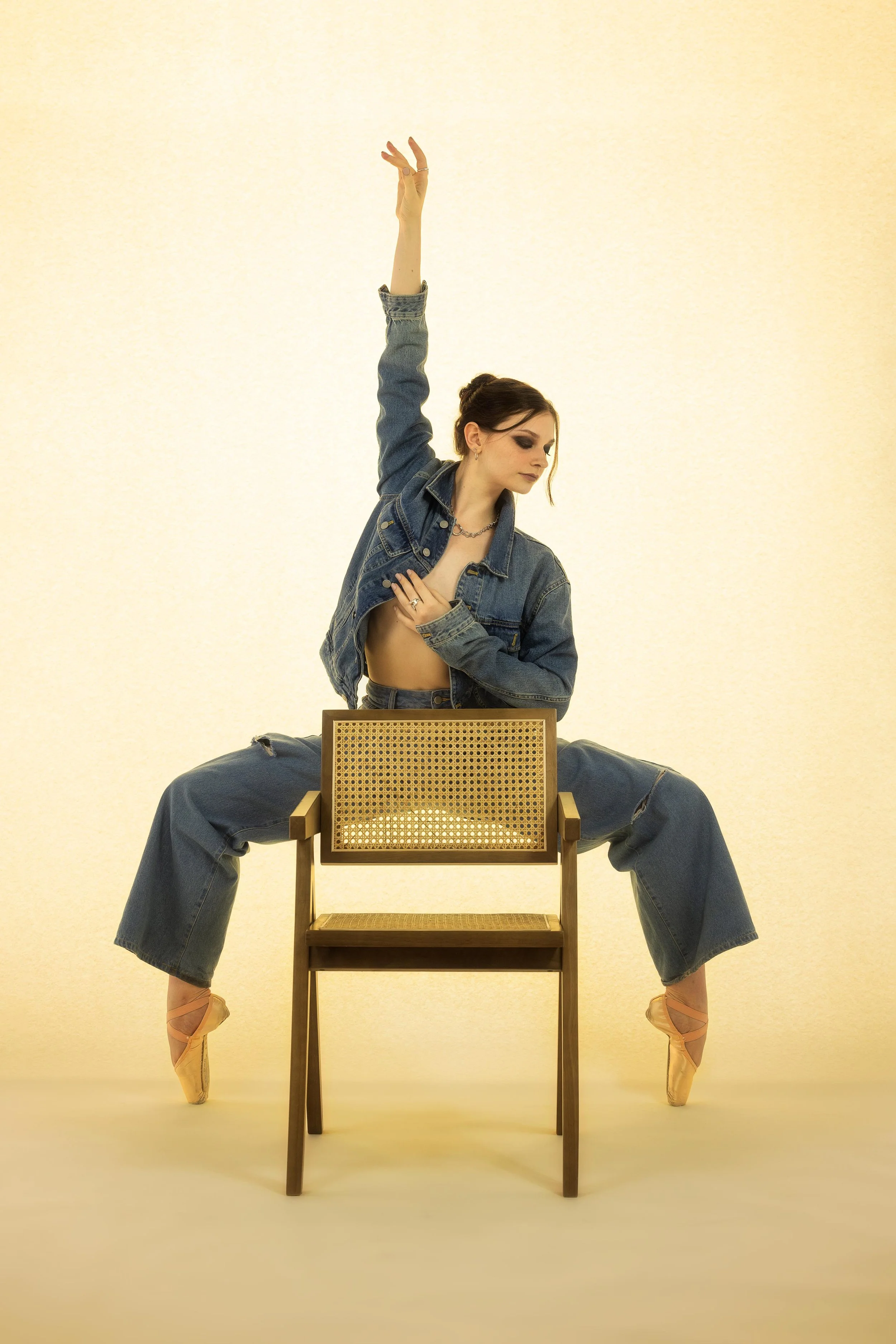 A dancer wearing a denim jacket and wide-leg jeans, performing a contemporary dance pose with one arm raised and the other resting on her chest, balancing on toes in pink ballet shoes, seated on a wooden chair in front of a plain light-colored backgr