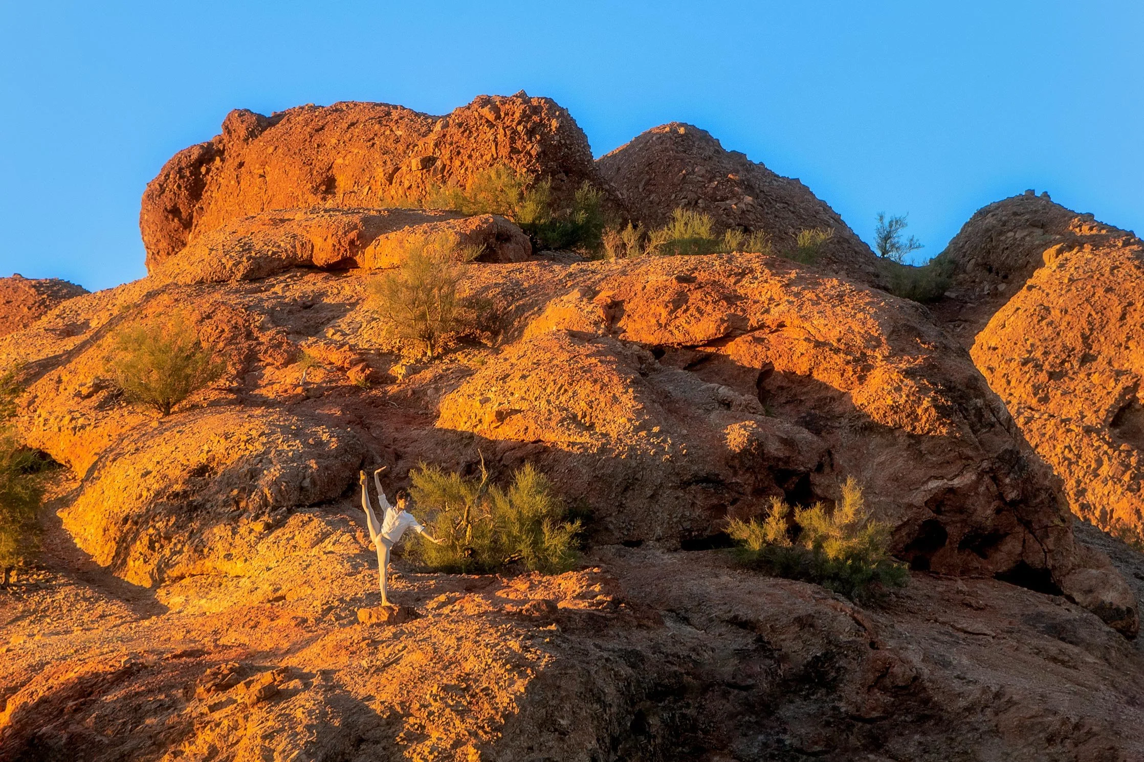 A person lifting the leg in a rocky, desert landscape with orange rock formations and sparse green vegetation, under a clear blue sky at sunset.