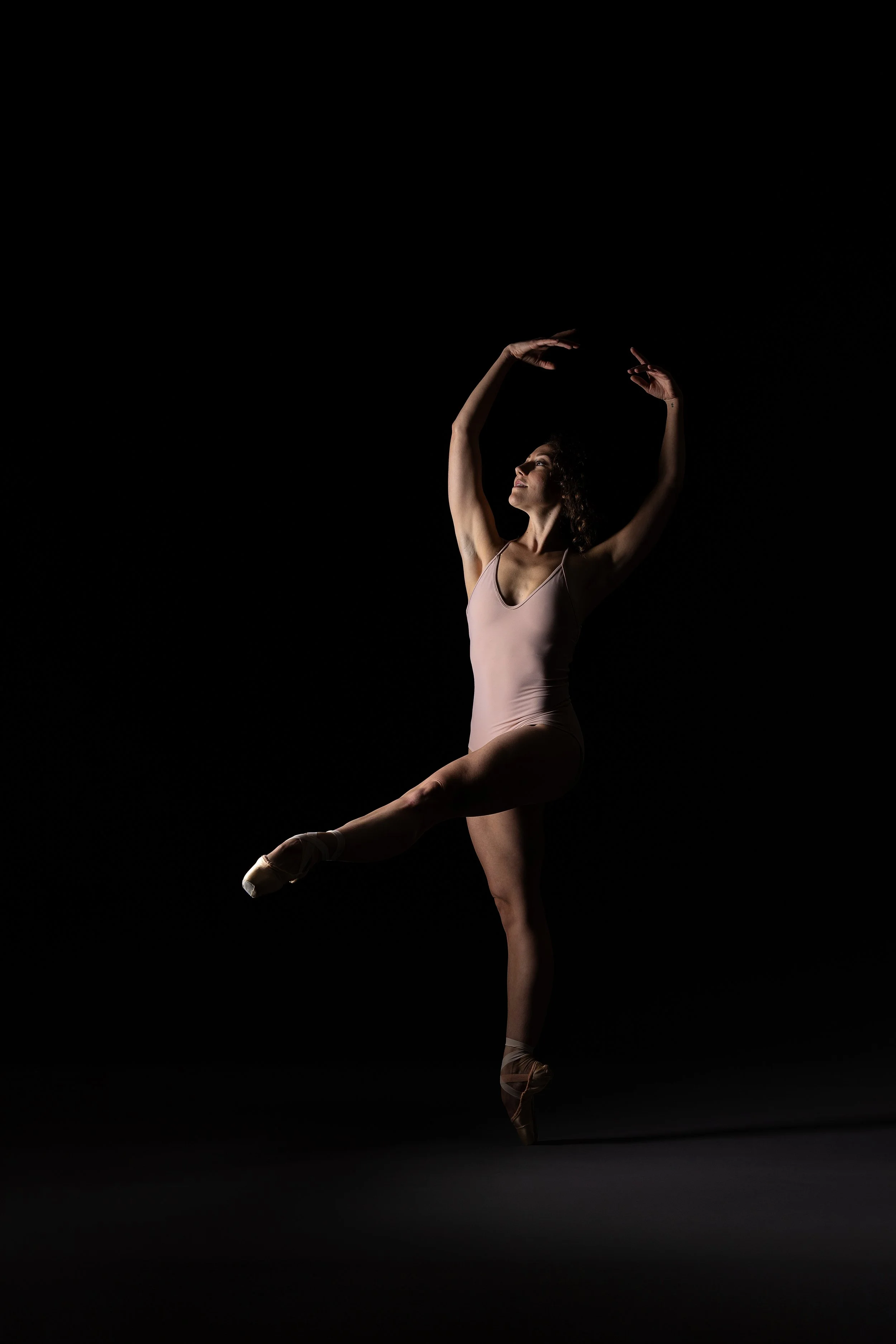 Ballet dancer in pink leotard performing a pose against a dark background.