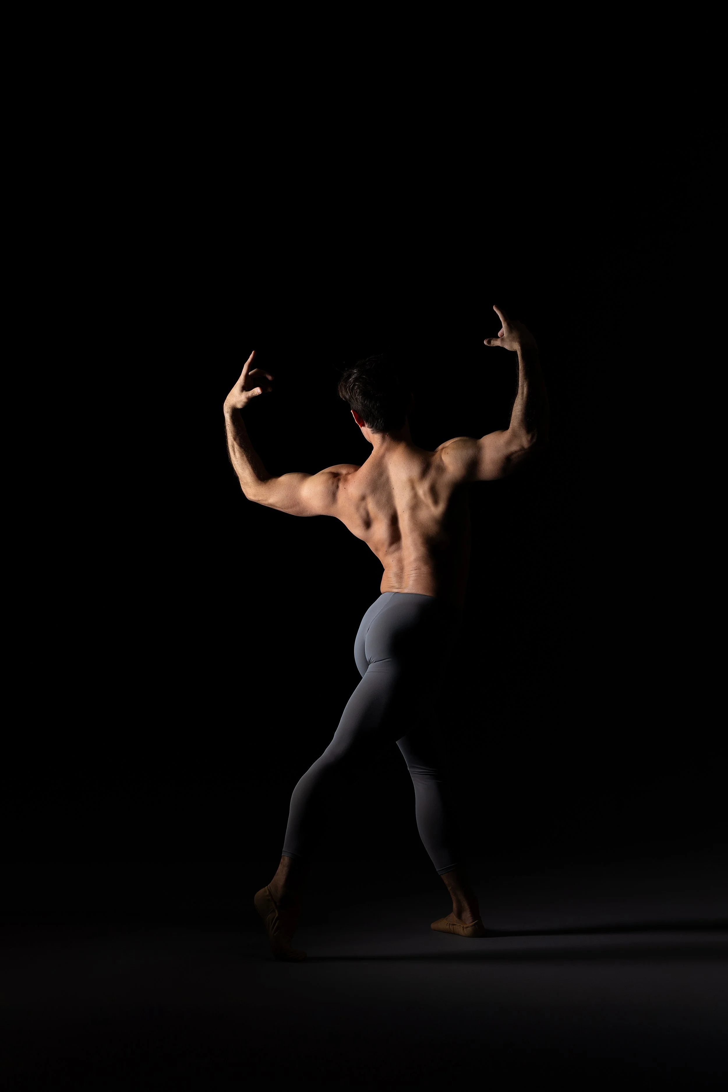 A shirtless man in dark pants and tan shoes is balancing with his back to the camera, flexing his muscles against a black background.