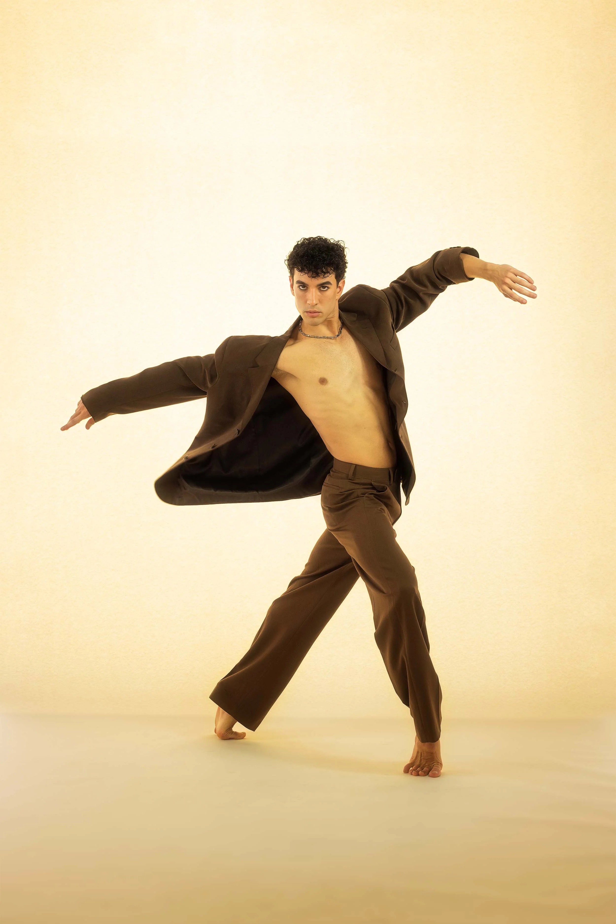 A man dancing in minimalist setting with beige background, wearing brown pants and a dark blazer, with curly black hair and a chain necklace, in a dynamic pose with arms extended and one leg bent.
