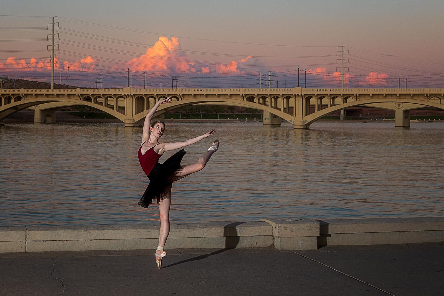 A ballerina in a red top and black tutu performing a dance pose on a paved surface by a river during sunset, with a bridge and electrical towers in the background and pink clouds in the sky.
