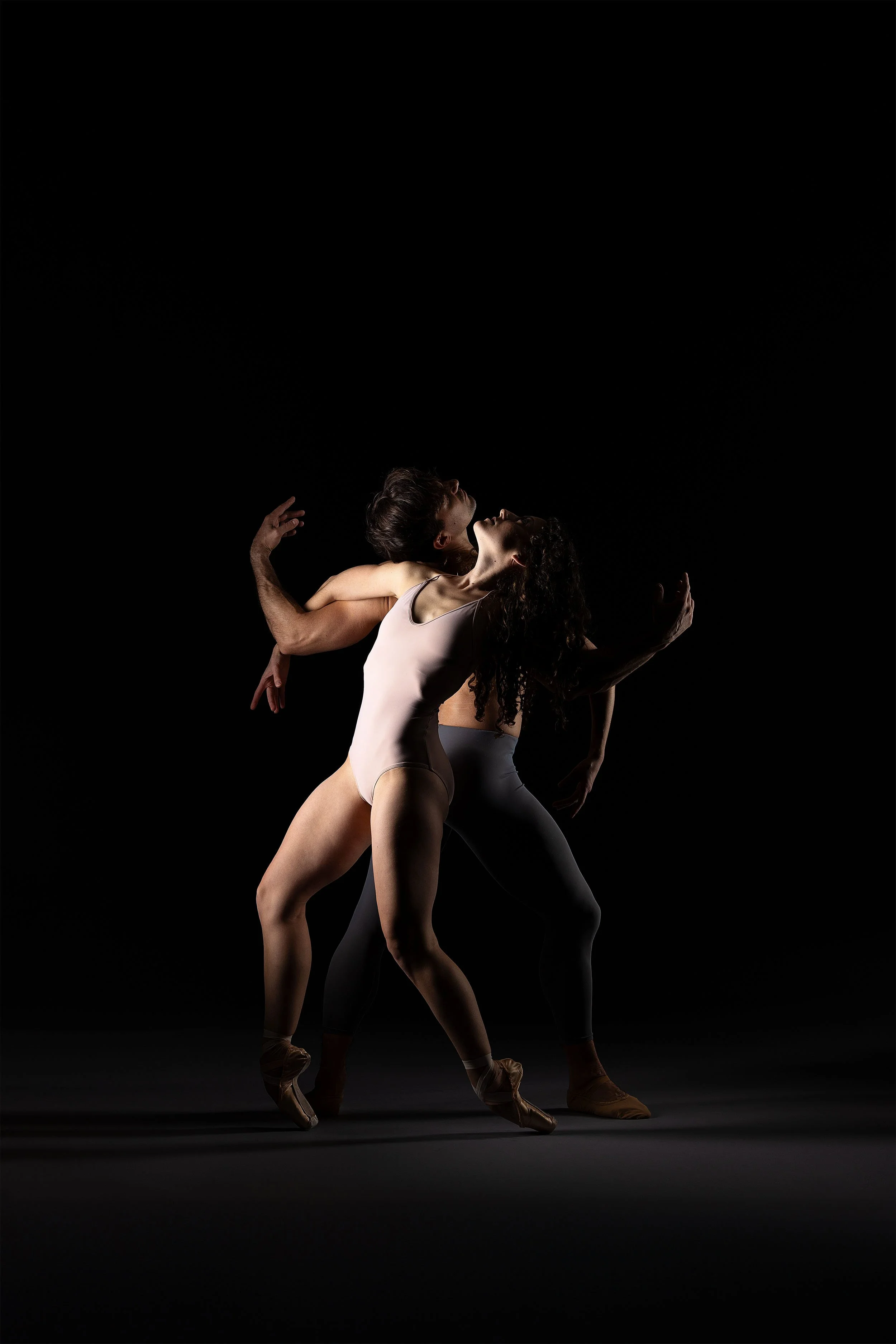 Two ballet dancers performing a dance move with a black background. One dancer is wearing a beige bodysuit, and the other is in gray leggings and a dark top.