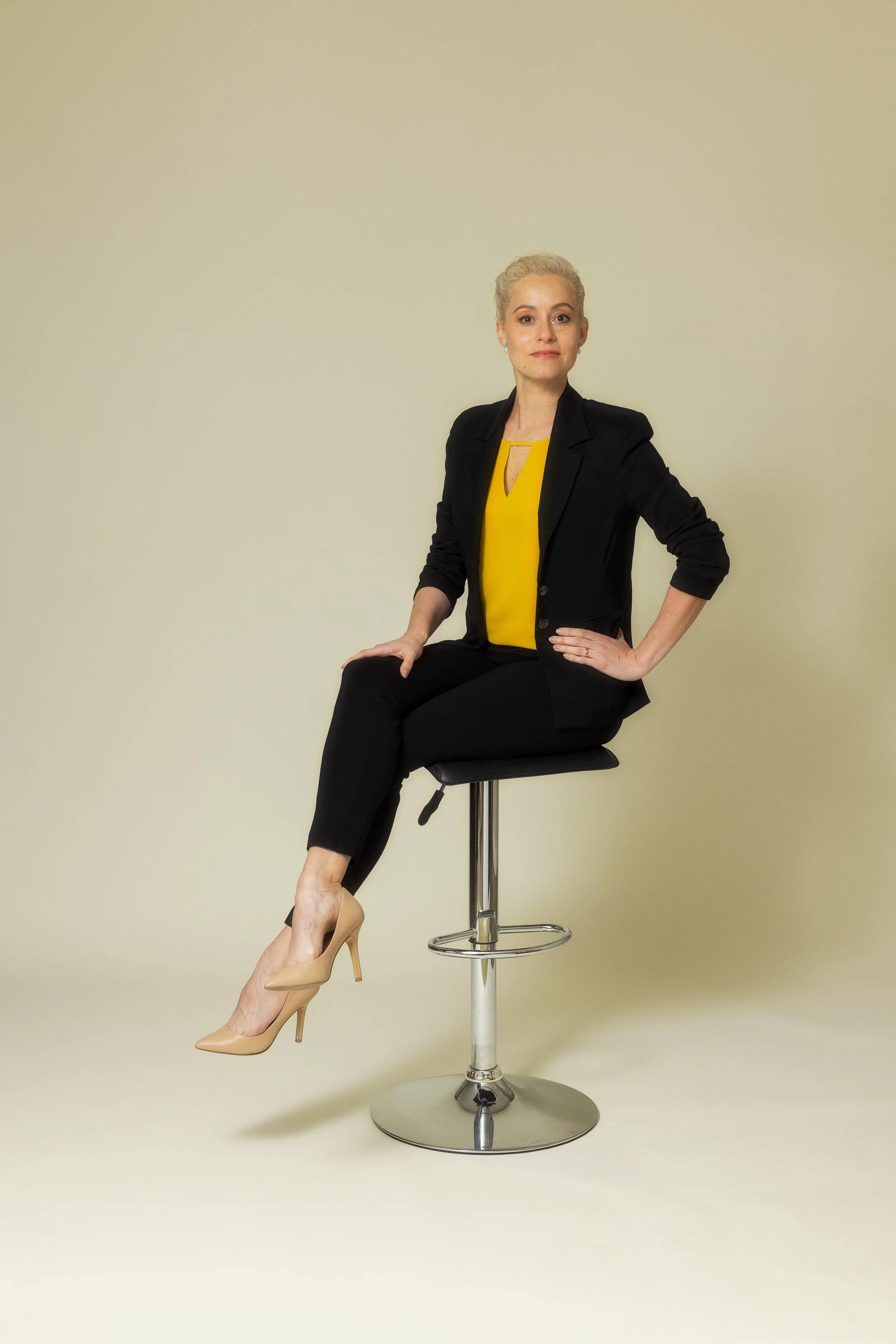 A woman with blonde hair sitting on a black stool, wearing a black blazer, black pants, beige high heels, and a yellow top, against a plain light background.