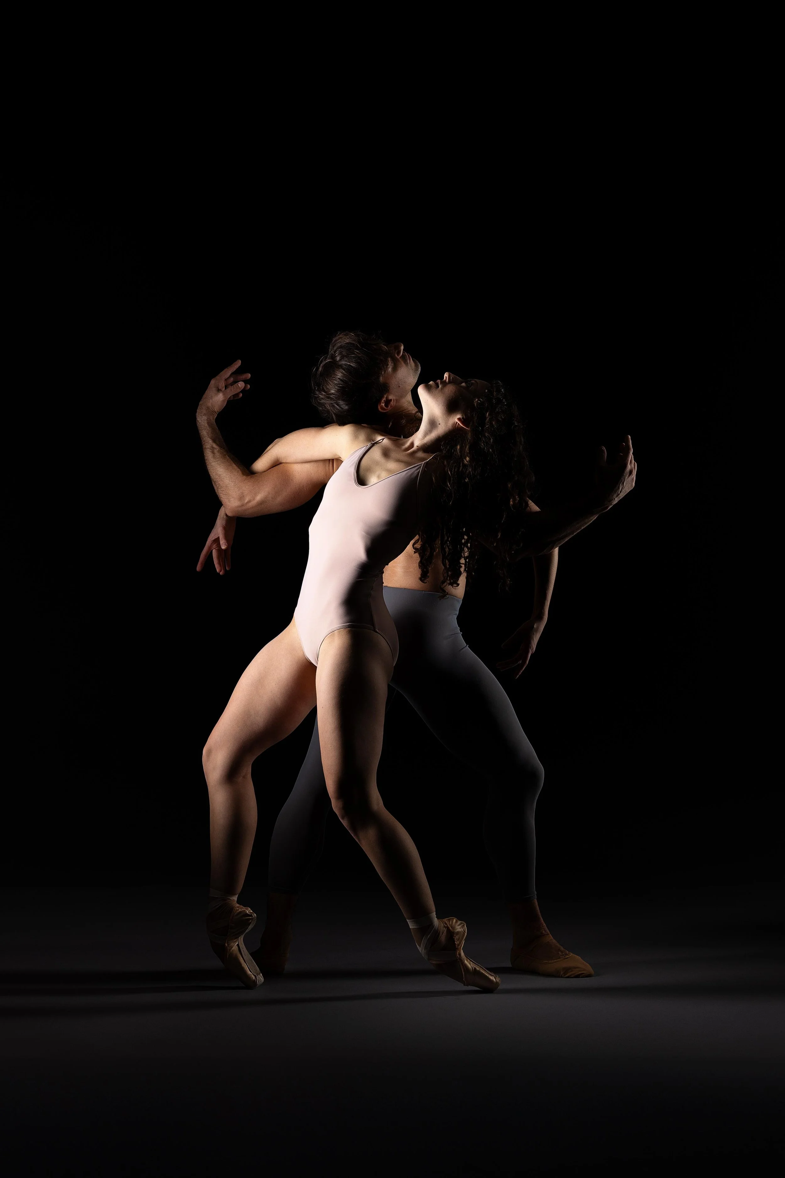 Two ballet dancers in a dramatic pose against a black background, with one dancer leaning back and the other supporting her, both in ballet attire and pointe shoes.