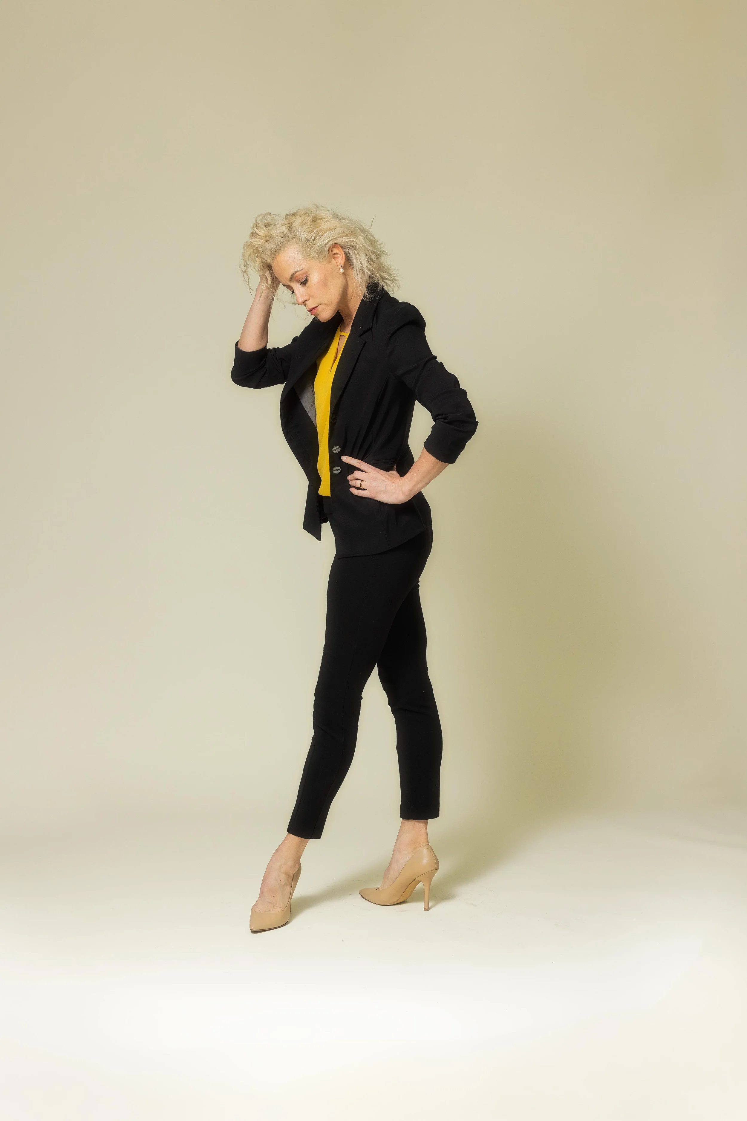 A woman with blond, curly hair poses with one hand on her head and the other on her hip, wearing a black blazer, yellow blouse, black pants, and beige high heels, standing against a neutral background.