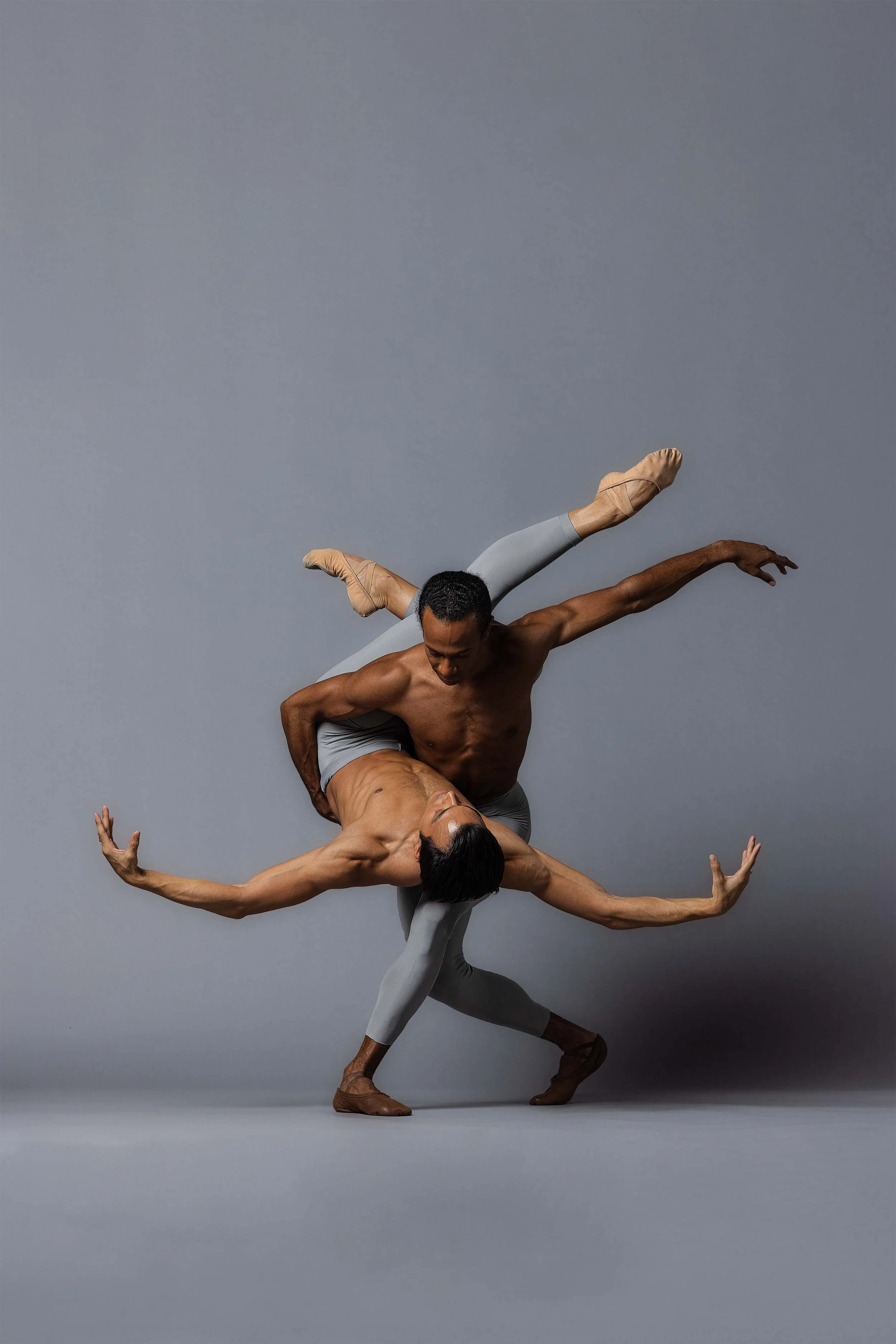 Two male ballet dancers with dark and light skin, performing an intricate pose against a plain gray background. One dancer pushes and supports the other as they balance with one leg extended behind them and arms reaching outward.