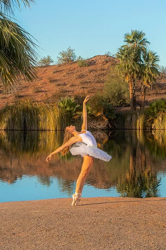 A ballet dancer in a white tutu practicing on the beach near a small body of water with palm trees and desert hills in the background.
