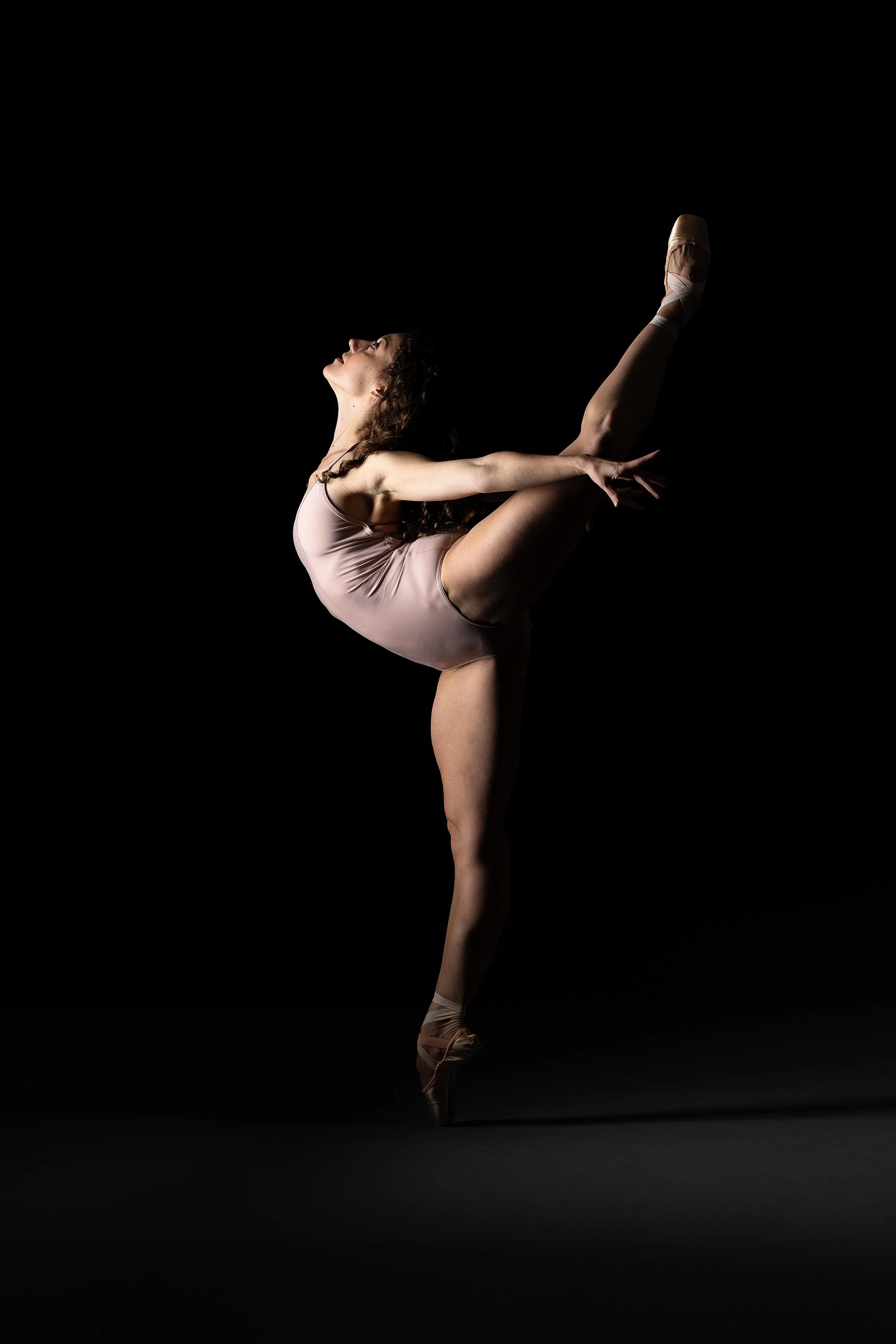 A ballerina performing a dance pose with one leg raised high and arms extended, against a black background.