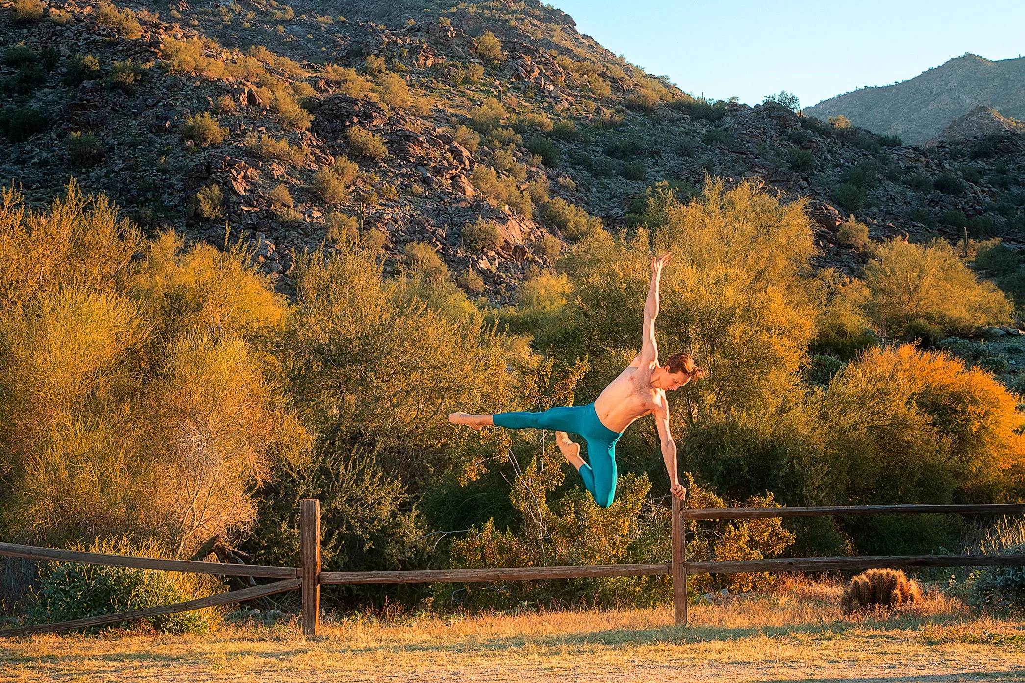 A male dancer practicing on a wooden fence in a desert landscape with yellow and orange trees and rocky mountains in the background during sunset.