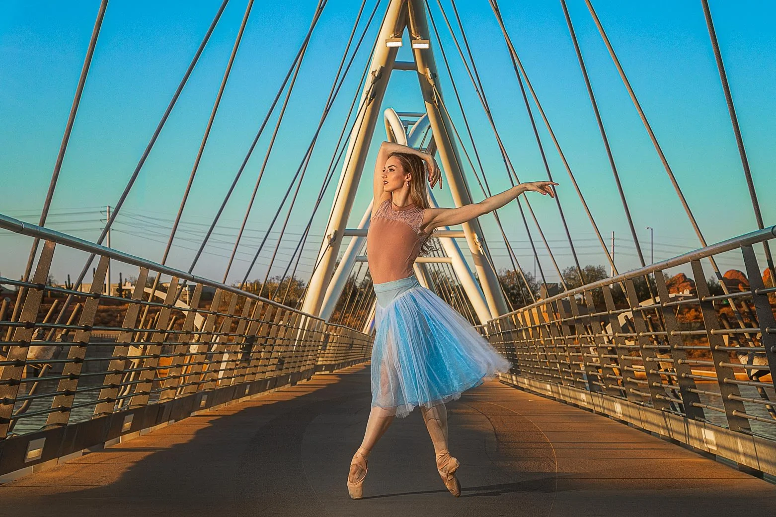Ballet dancer in a pastel-tinted tutu performing on a modern suspension bridge during sunset.