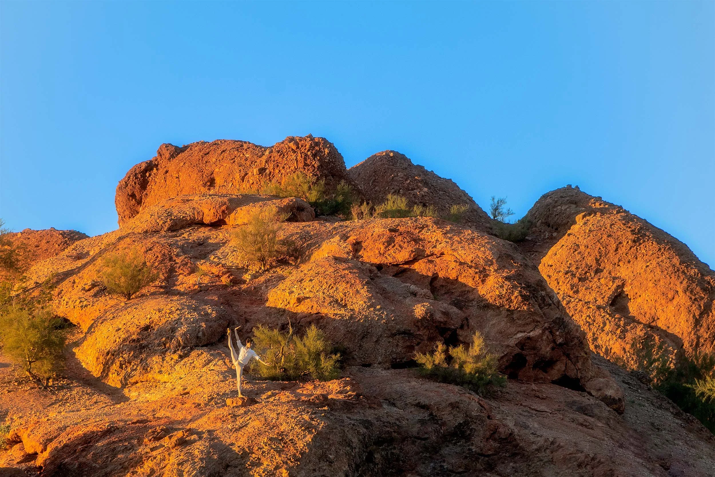 Desert landscape with rocky hills bathed in orange sunlight, sparse green bushes, and a clear blue sky.