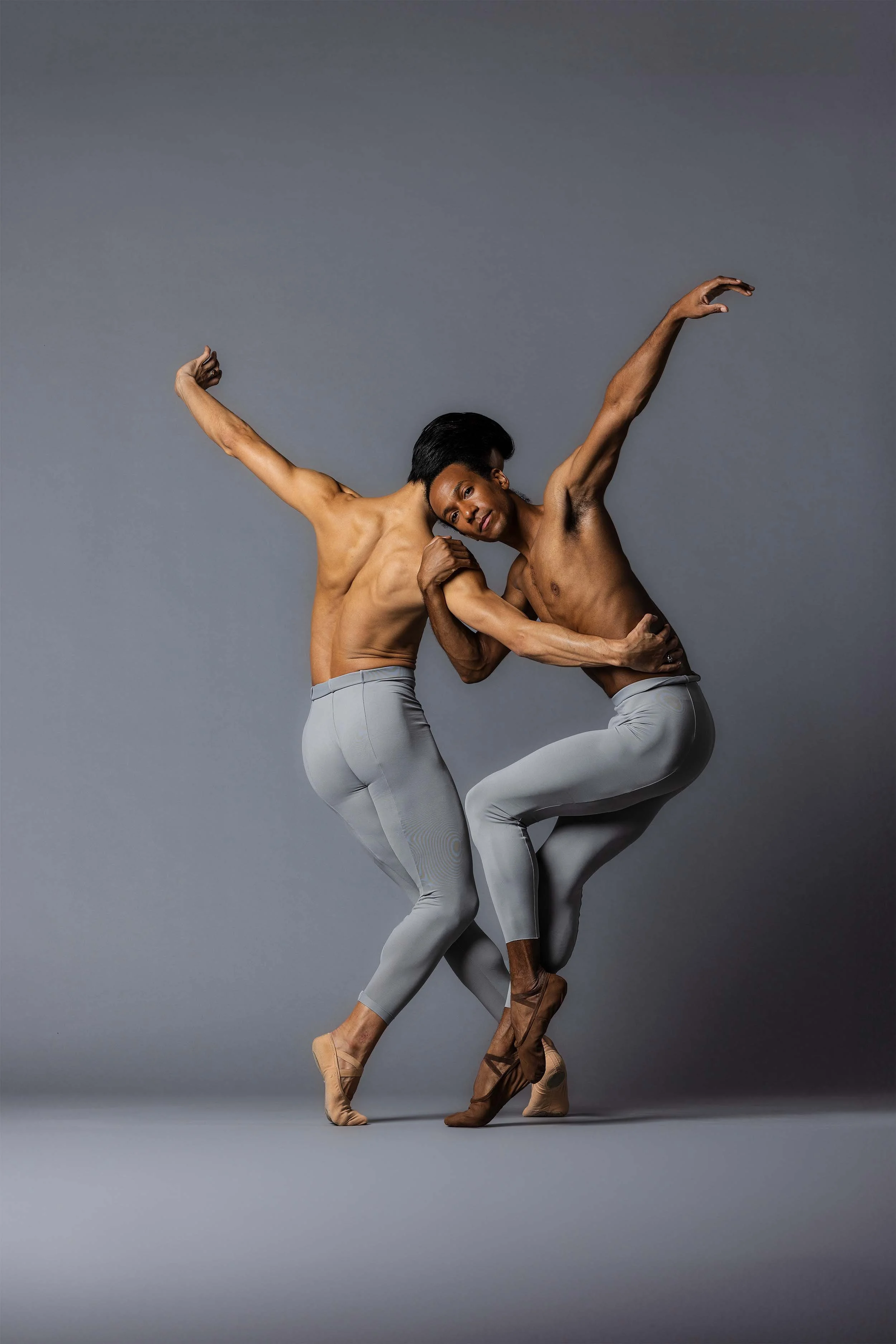 Two male ballet dancers with dark skin in gray tights and beige dance shoes perform a pose against a gray background. They are shirtless, with one dancer's head resting on the other's shoulder, both with arms raised in expressive positions.